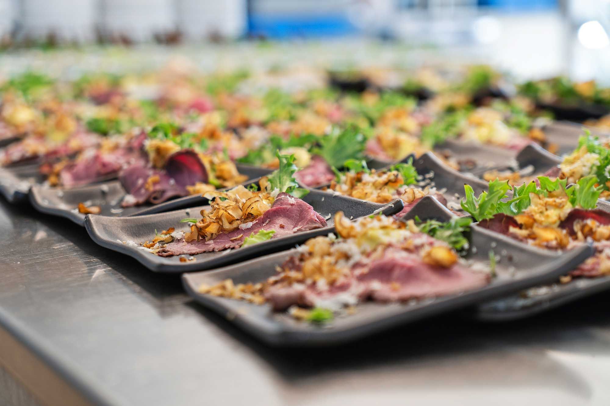 En lang række krydrede retter med rødt kød og grønne salatblade, der er udsat på en metaloverflade i et køkken