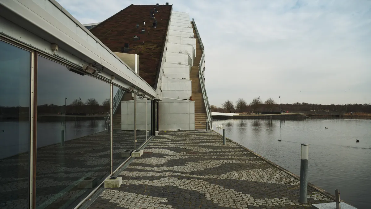 Bygning med grøn tagdækning og glastæppet facade vender ud mod vand. Moderne design med skrånende tag. Solrefleks på glas og fuglene svømmer på søen