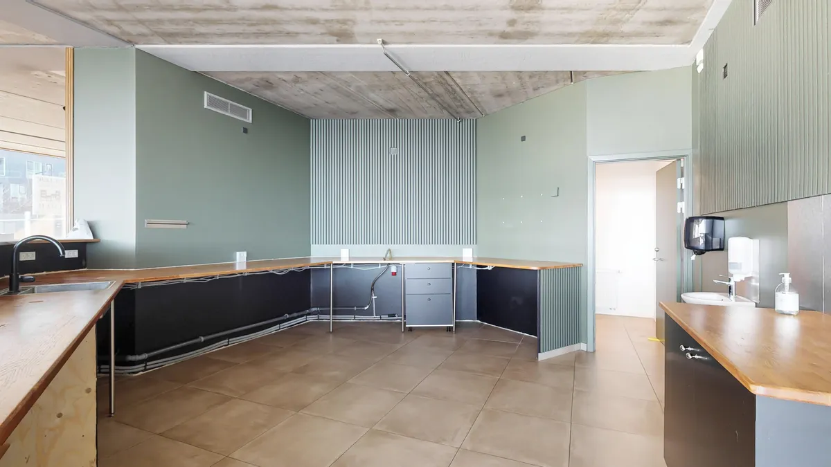 To tømmerkøkkener med højloftede lofter, lyse vægge og lyse gulve. To håndvask med vandhaner og en hånddesinfektionsdispenser.