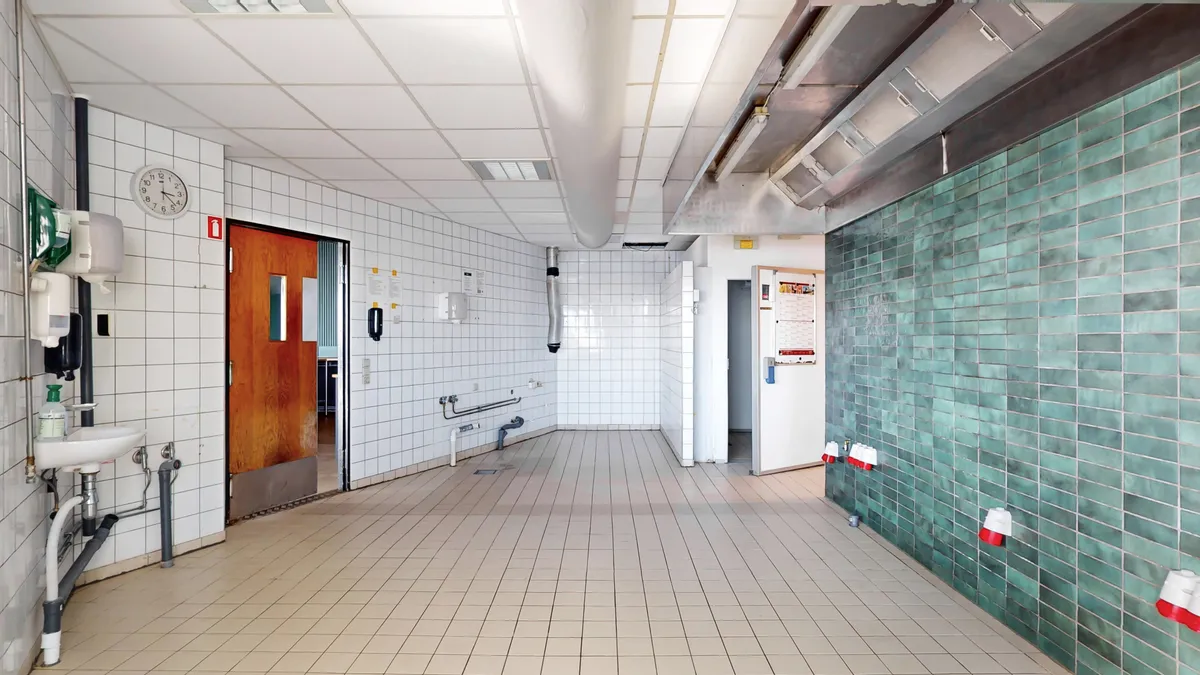 En tom toiletlokale med lyse, turkise og hvide keramiske fliser og en hvidt keramikskål med håndvask, hånddesinficering og toiletpapirautomat.