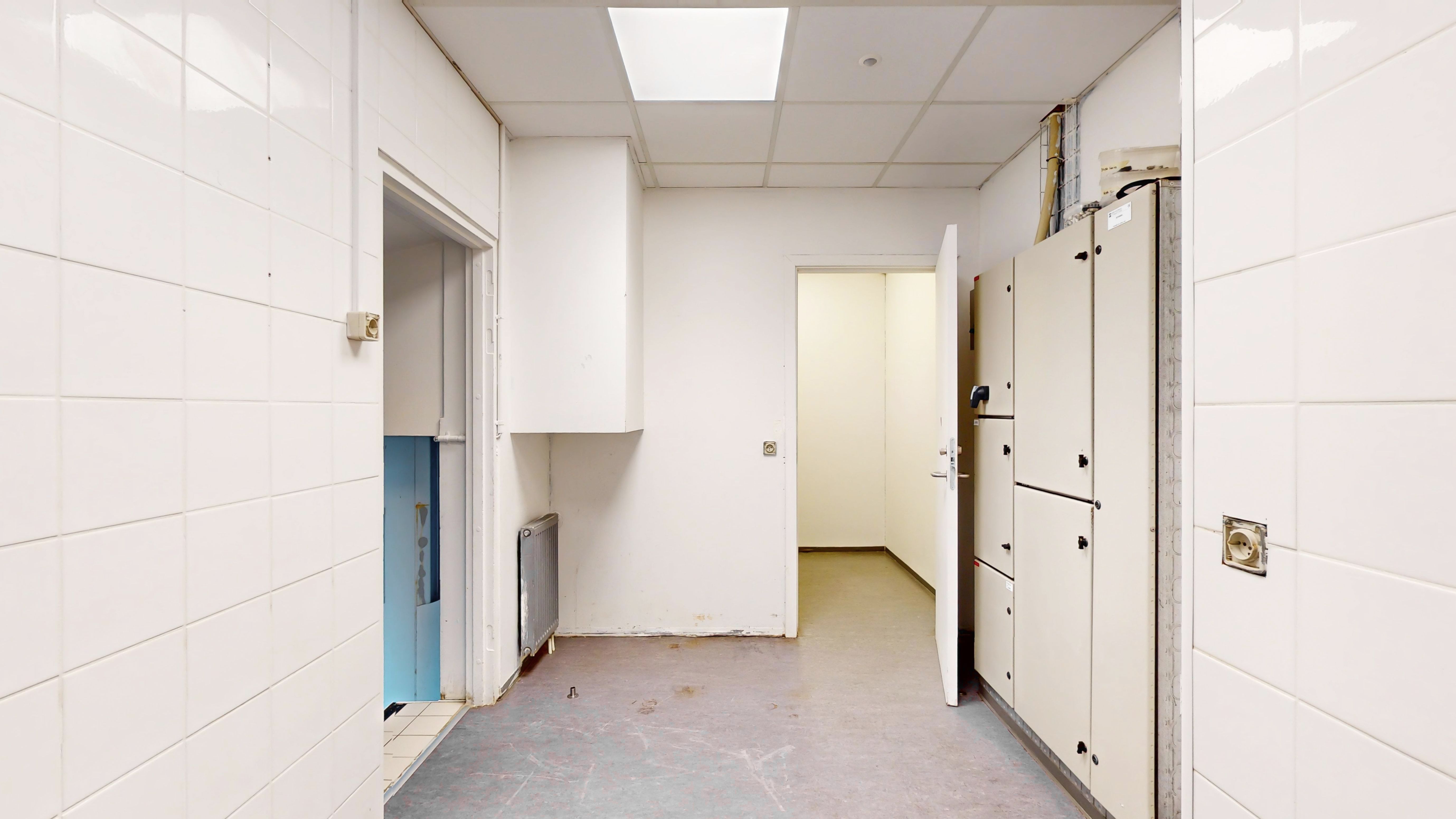 En stor rummelig og lys hall med hvide vægge og loft, åbne døre og kabinetter samt radiatorer og elektriske stik.