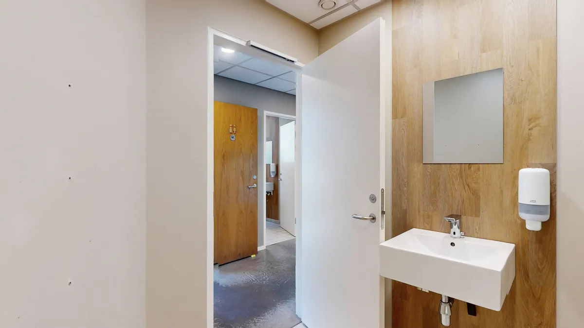 Rent hvidt badeværelse med lyse trævægge og hvidt toilet. Hvid sænk og håndvask med sæbeautomat og spejl. Dør med wc-skilt.
