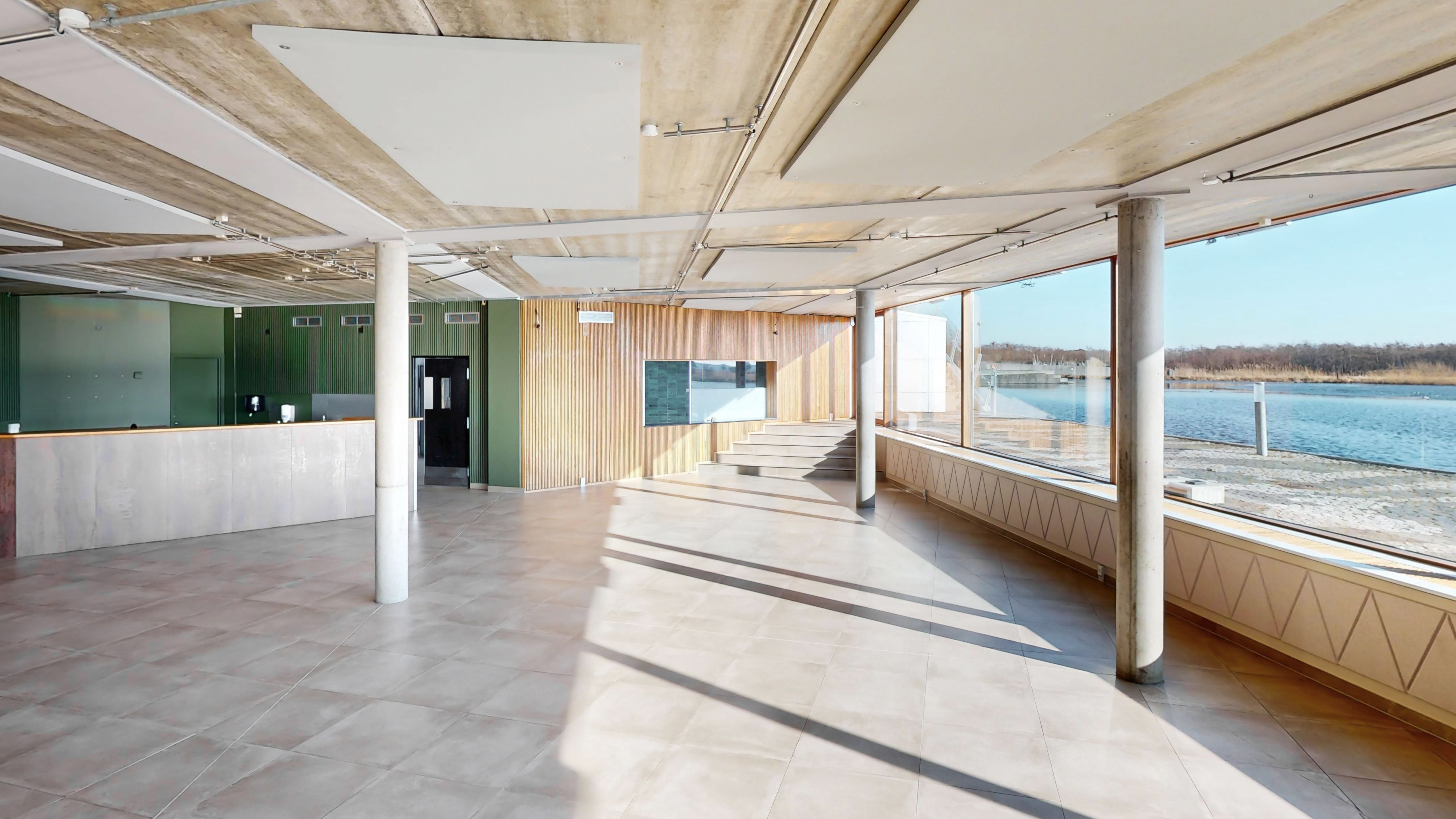 Her ses et tømt rum med terrasse og udsigt til vandet og skov. Loftet og væggene er beklædt med lyse paneler og gulvbelægningen er lysegrå.