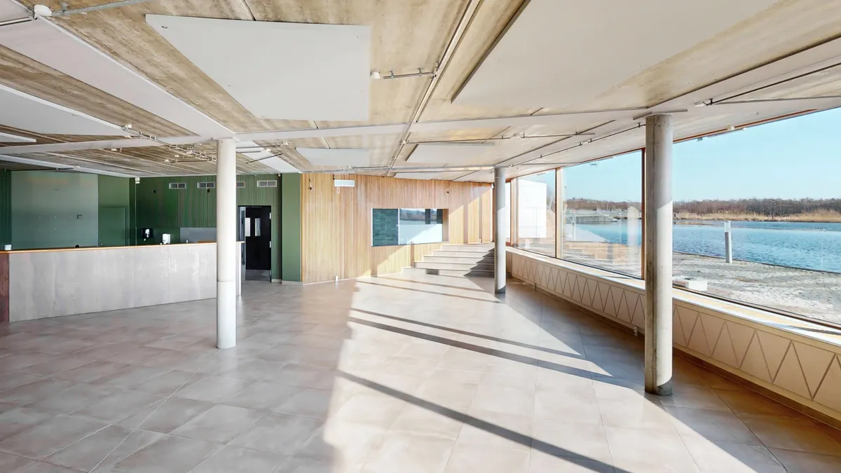 Her ses et tømt rum med terrasse og udsigt til vandet og skov. Loftet og væggene er beklædt med lyse paneler og gulvbelægningen er lysegrå.