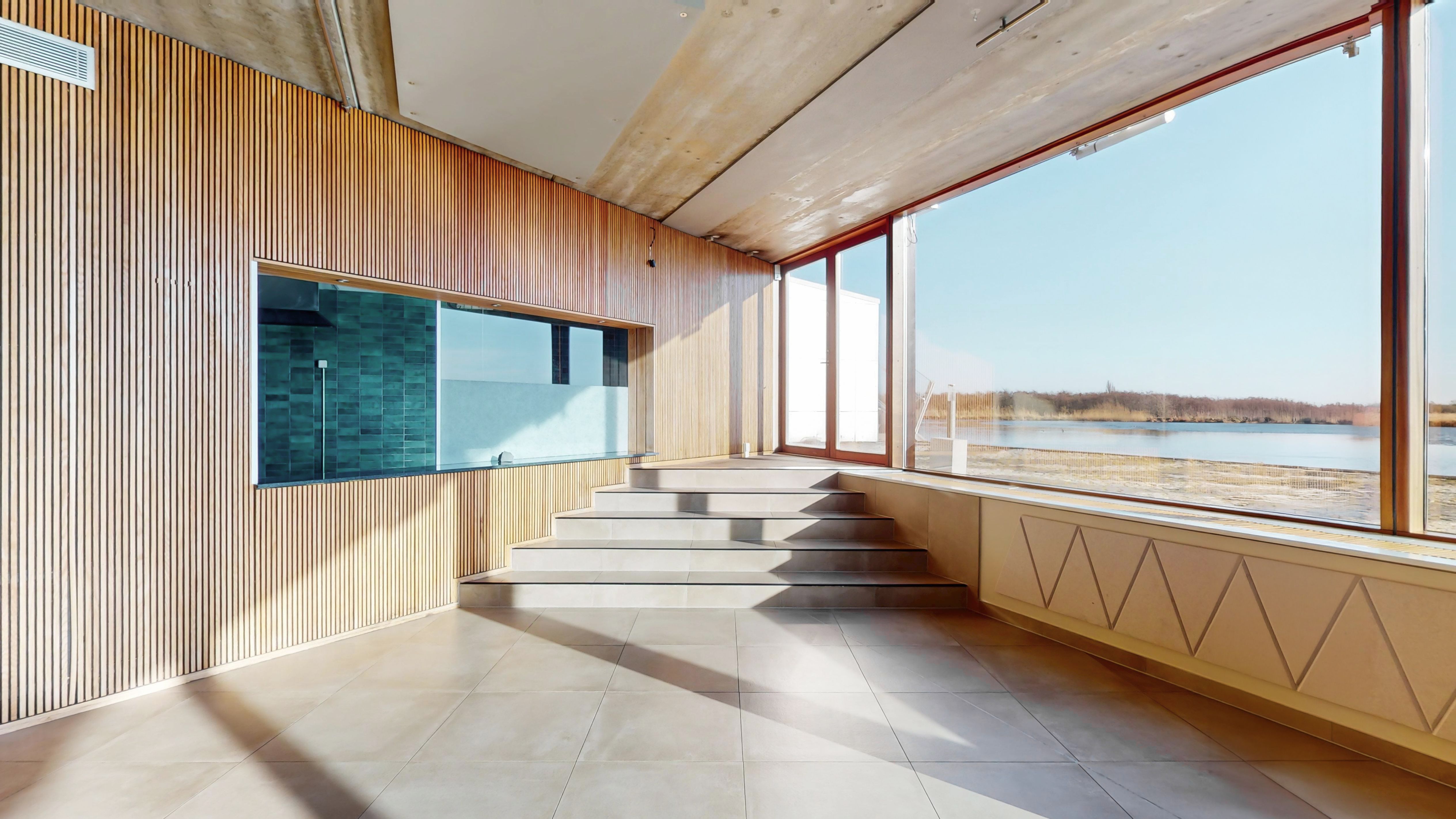 Lys og rummeligt rum med gult gulv og mønstret træbeklædning, trapper og vinduer med udsigt over vandet og landskabet