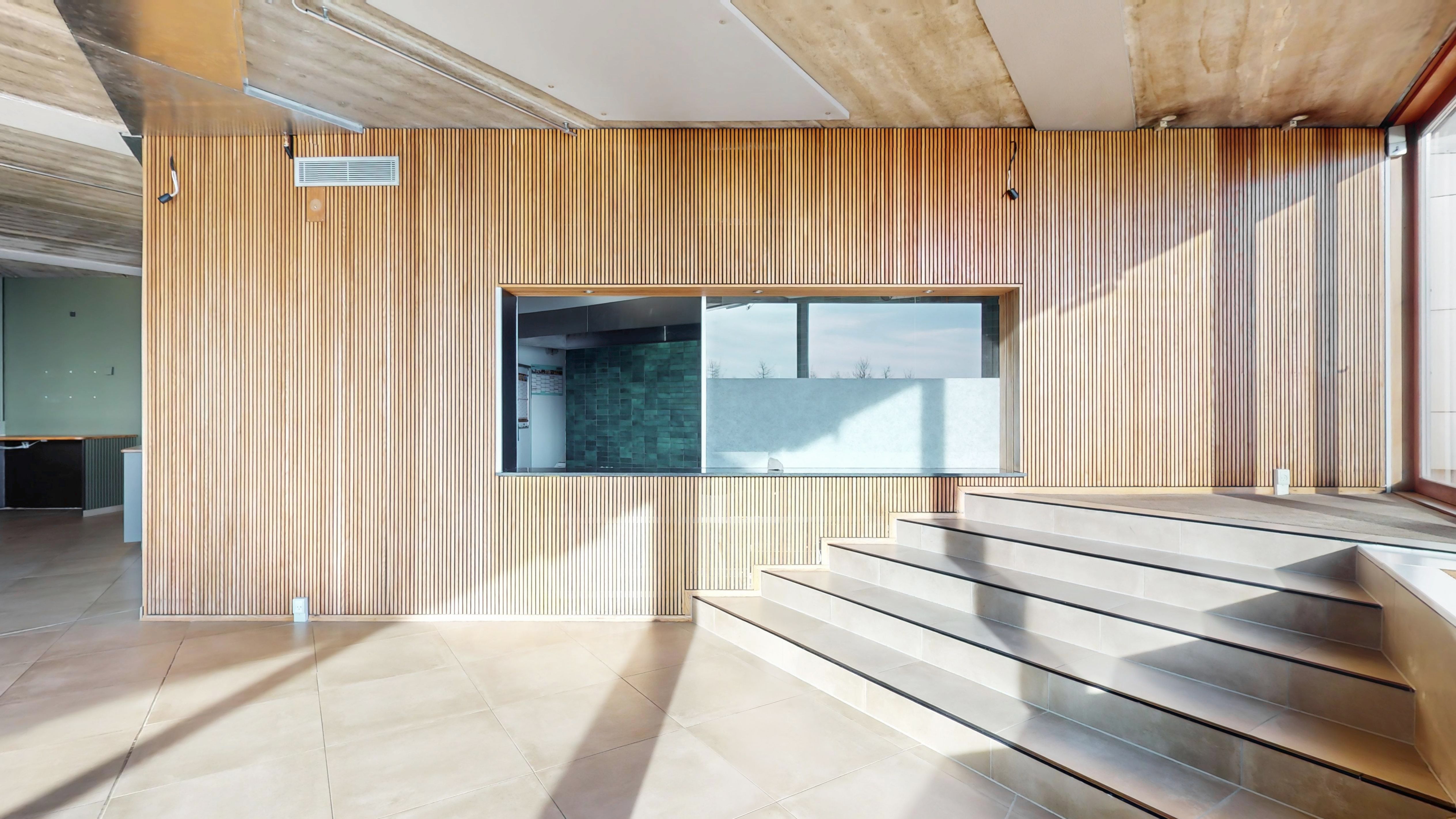 Lys indlysende værelse med gulv og træbeklædte vægge, indre glasvindue med udsigt til udendørs arealer, og stige med lysegult gulv