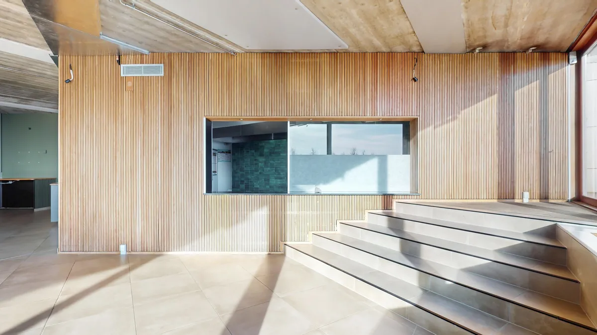 Lys indlysende værelse med gulv og træbeklædte vægge, indre glasvindue med udsigt til udendørs arealer, og stige med lysegult gulv