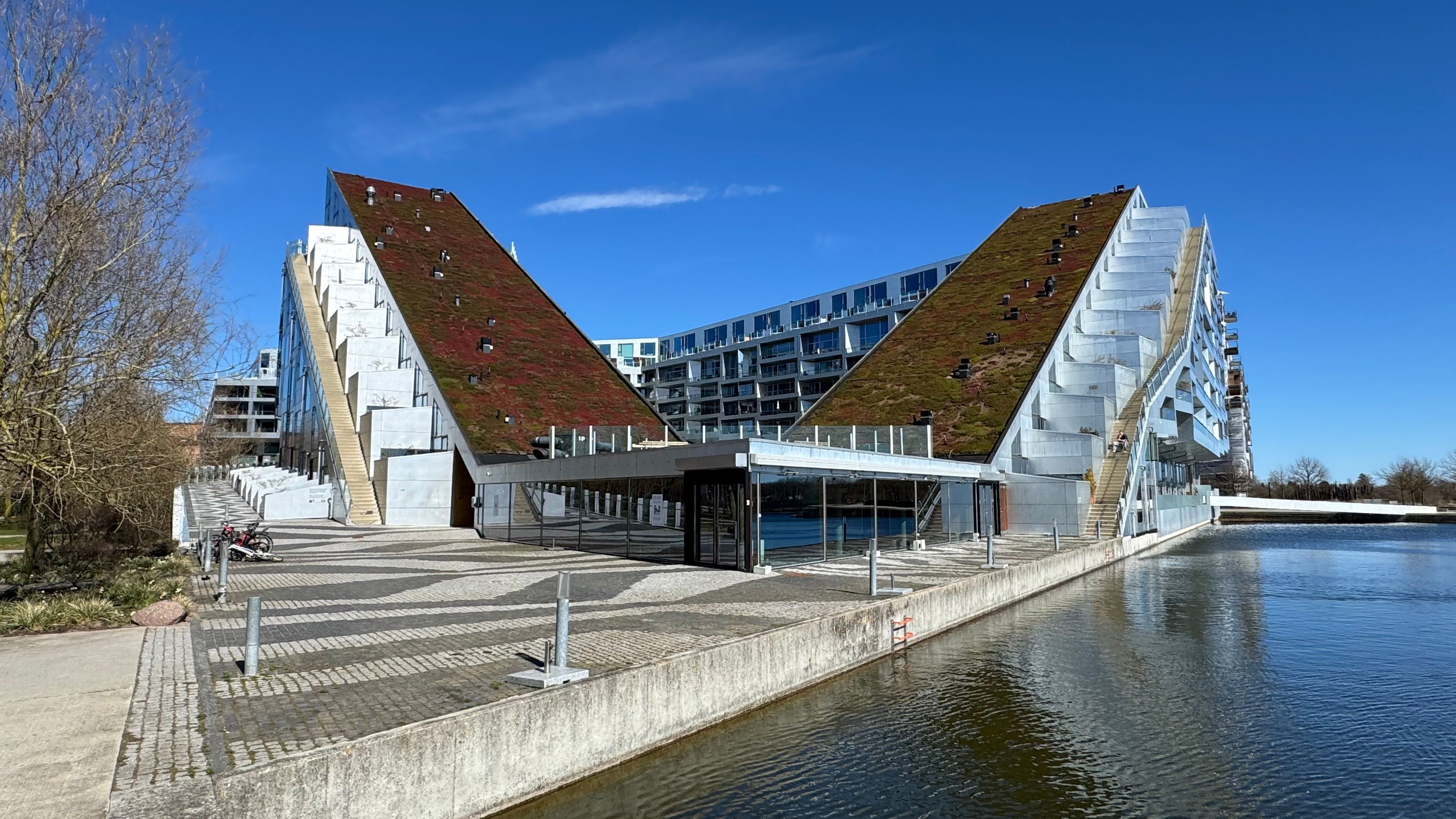 Moderne bygning med grønne tag ved en kanal i København S. En cykel parkeret og en terrasse med glasoverdækning