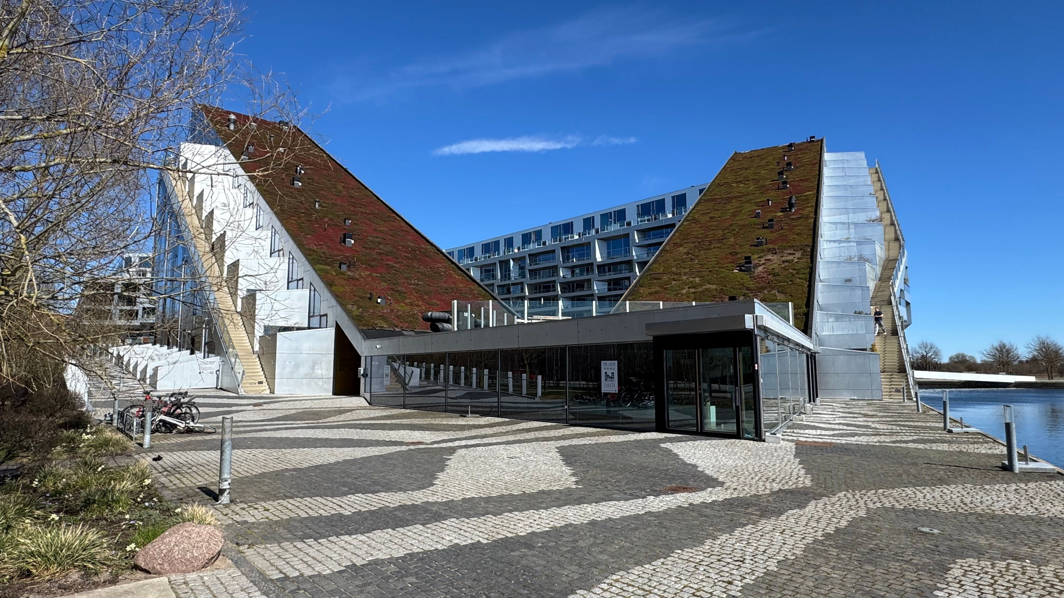 Moderne bygning med grøn tagdækning og glasvægge placeret ved vandkanten. Adgangsdør og terrasse med udsigt over vandet. Parkerede cykler og skulpturer langs gaden.