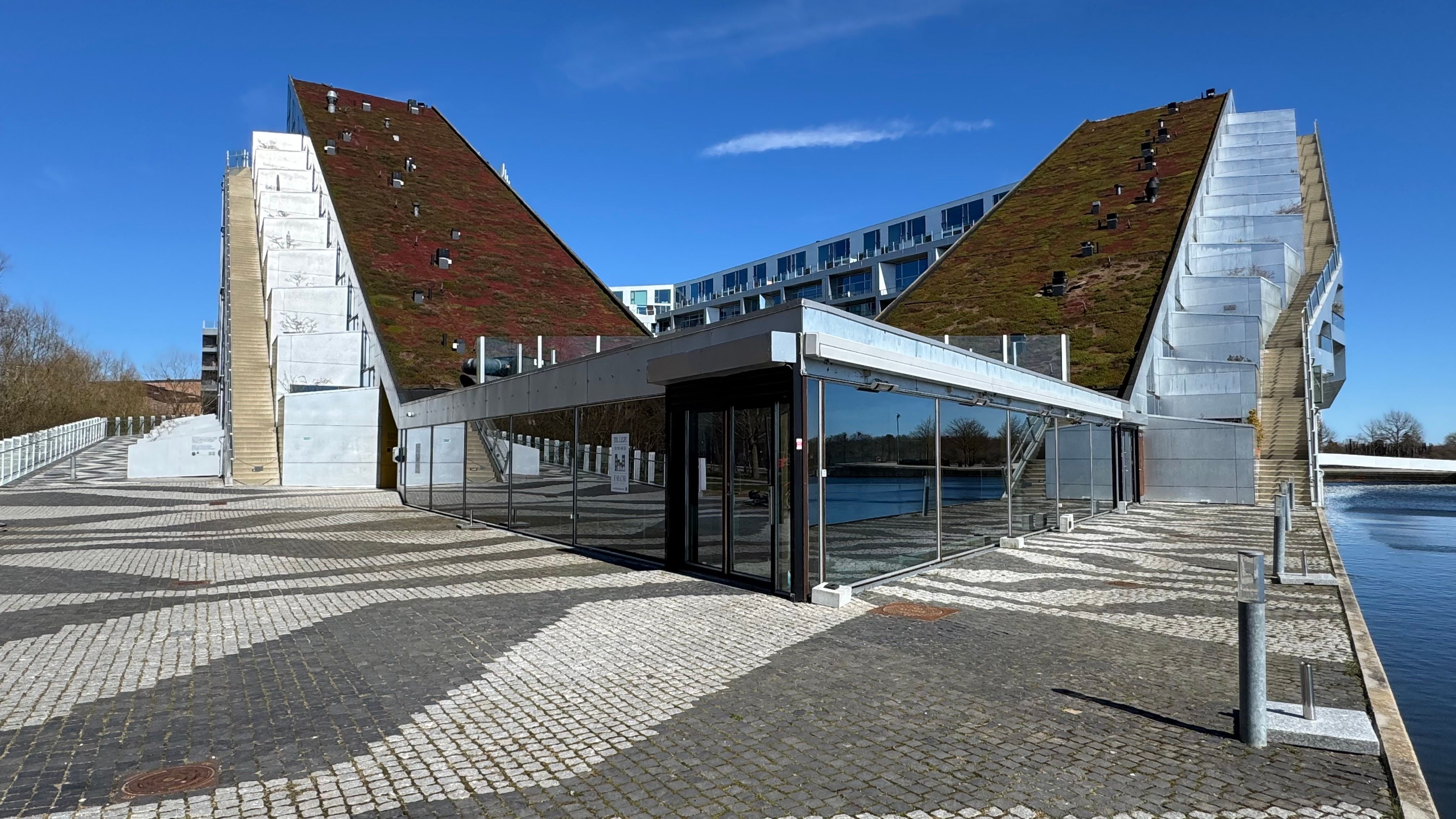 En moderne bygning med grønne tagdækning og glas facader ligger ved vandet med udsigt over en park.