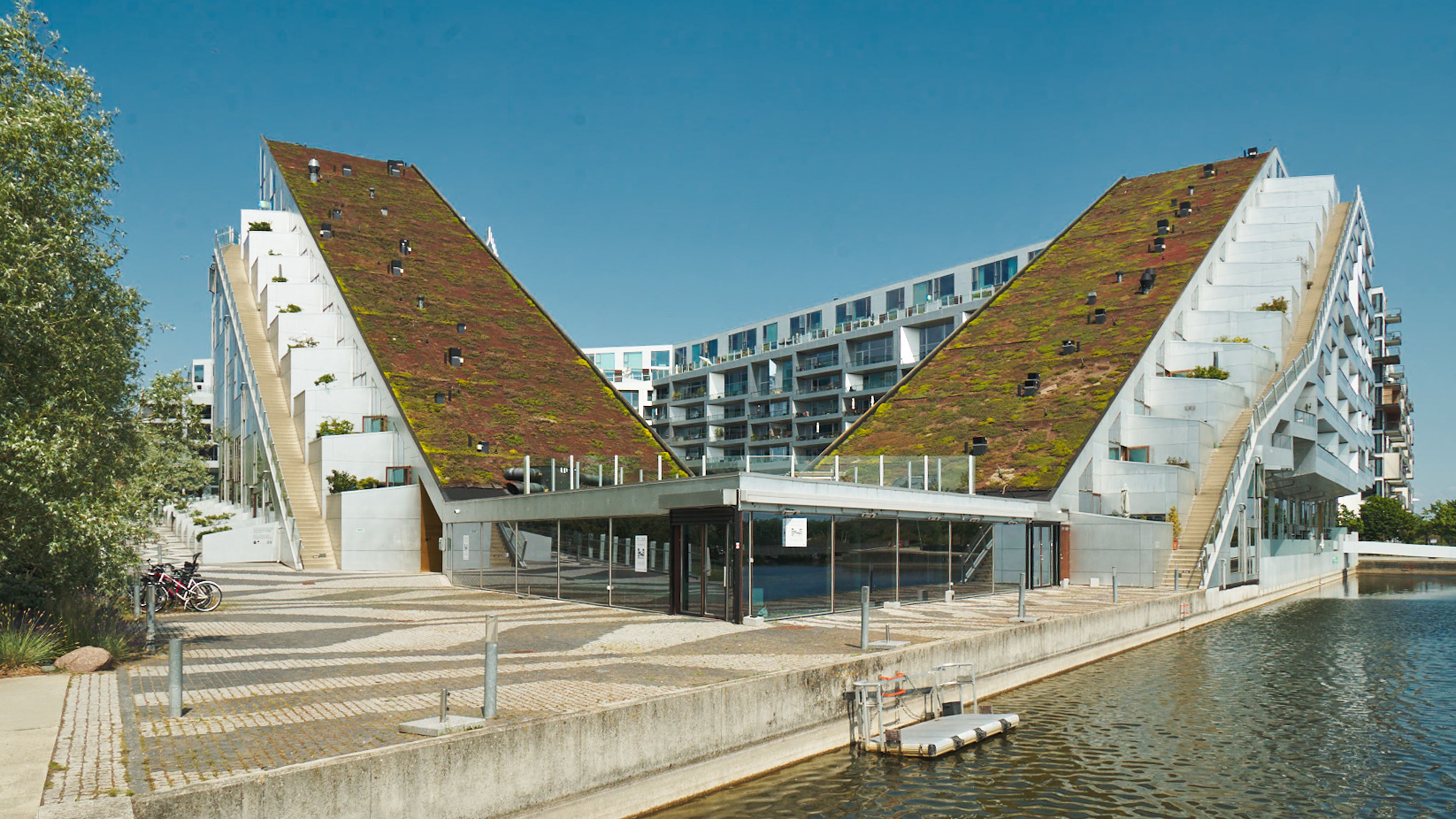 Moderne bygning med grønne tag i Ørestad. Glasvinduer og trapper. Vand og båd på kaj. Solrigt og indbydende område.