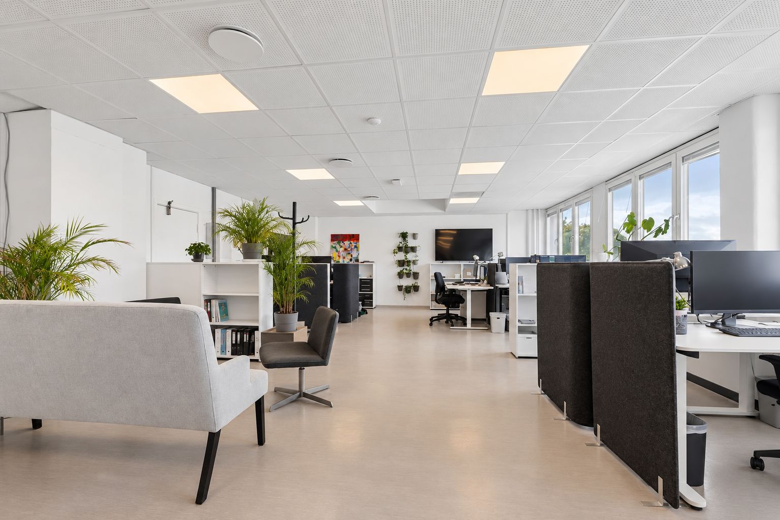 Et rektangulært, veludstyret kontor med høje hvide vægge, store vinduer og moderne møbler samt planter. To små sofaer, skriveborde, højtalere og flere skriveborde.