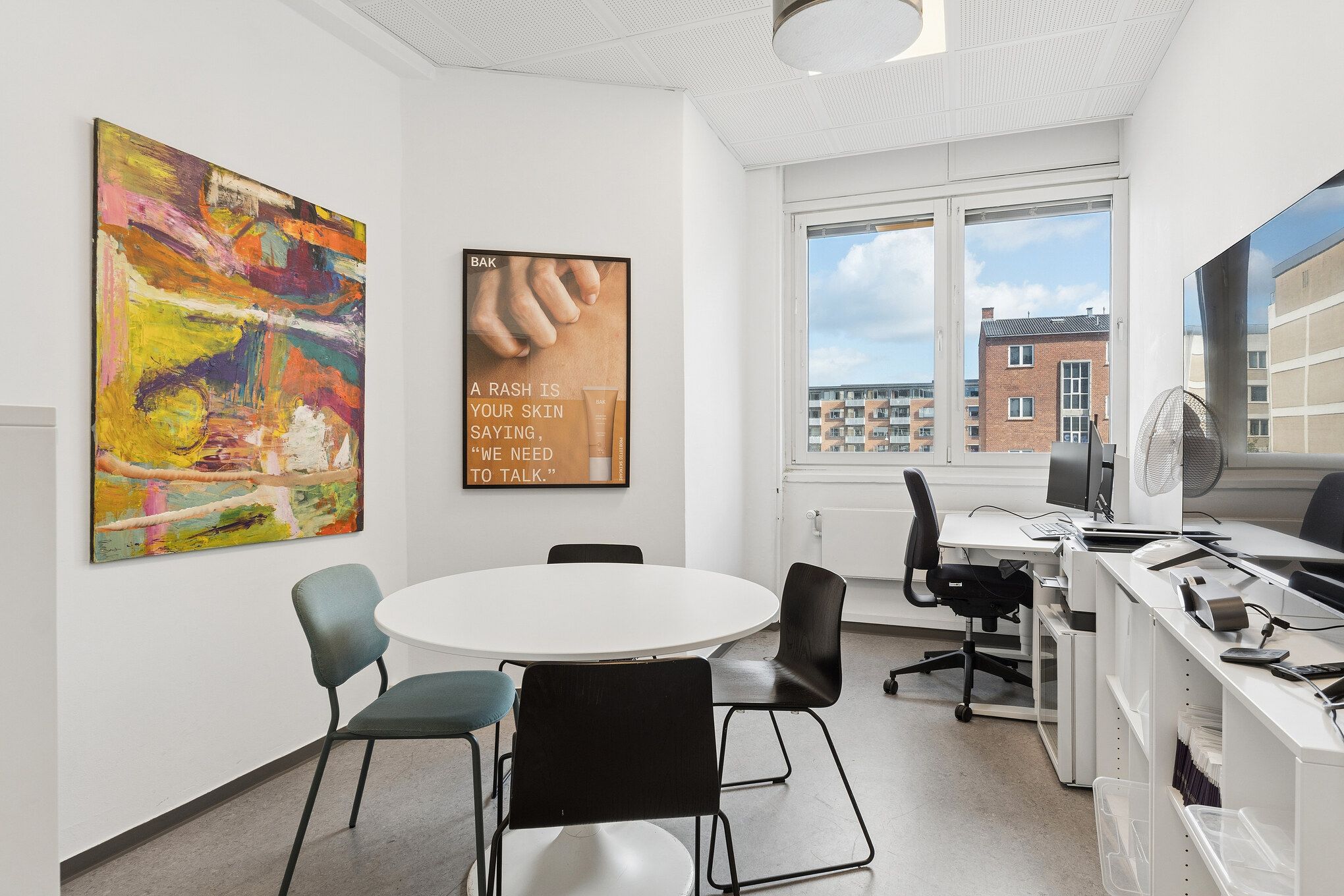 Et moderne kontor med rundt bord, fire stole, skrivebord med computer, stå- og skrifttegning, ståfan, kunstværk og vinduer med udsigt til bygninger og himmel.
