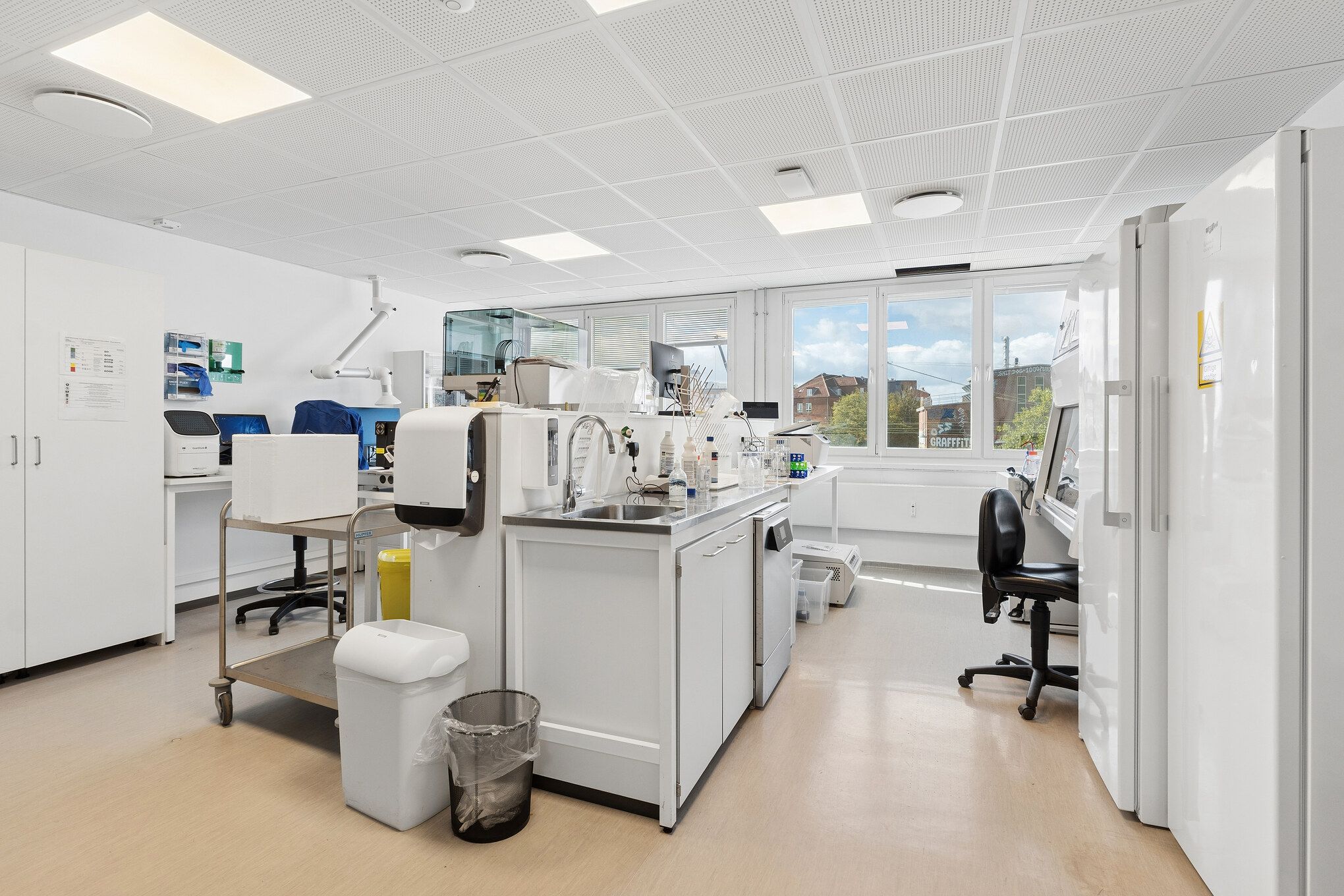 Laboratorie med laboratorieudstyr, højteknologisk udstyr, lysekroner, skarpe linjer, lyse gulve, høje vinduer, forskellige skuffer, stol og håndsæbeautomater samt et køleskab med advarselsskilt