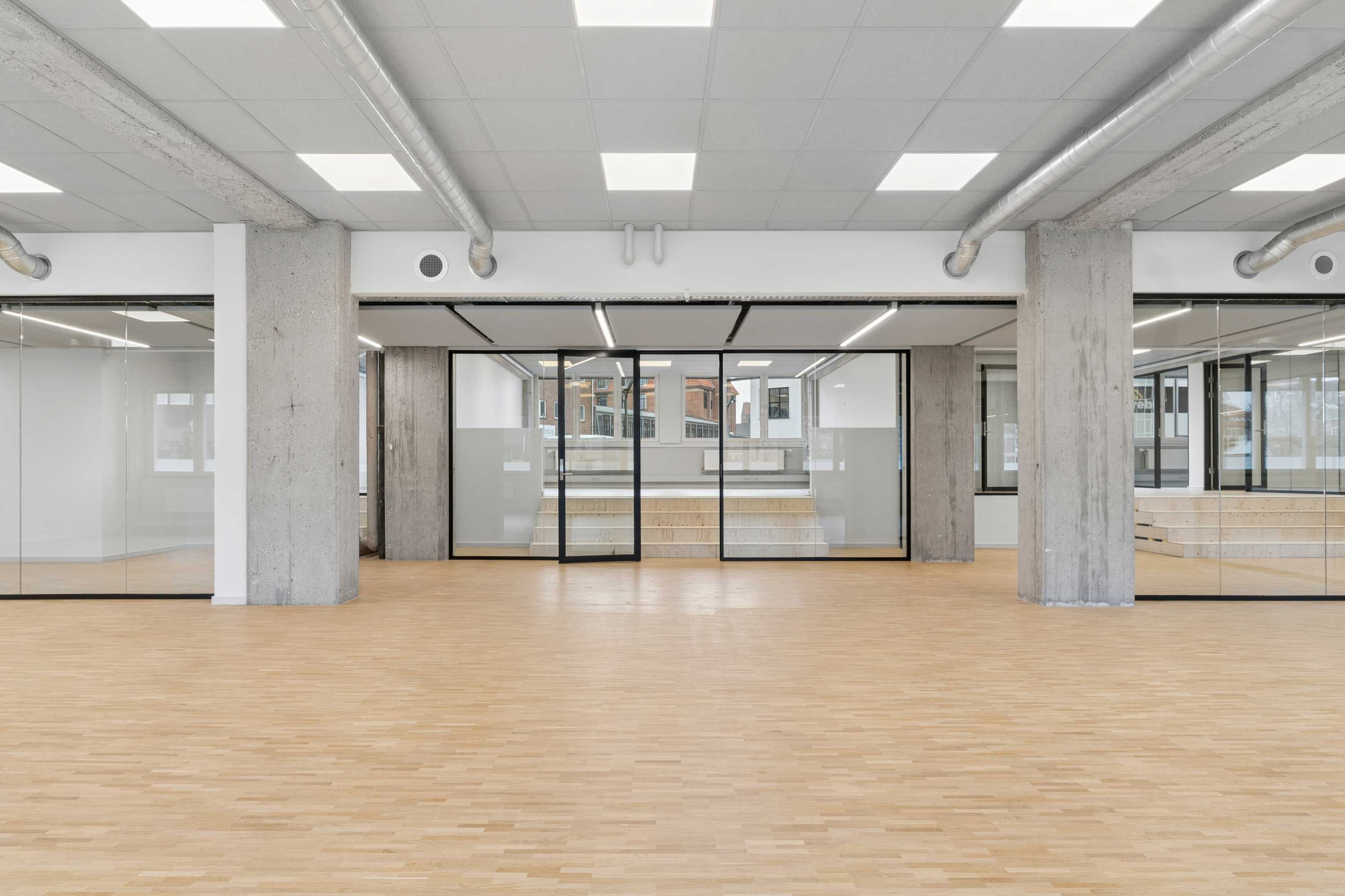 Et moderne kontor med store vinduer og lyse lofter samt trægulv. Større glasdøre med mørkere rammer og hvidt interiør.
