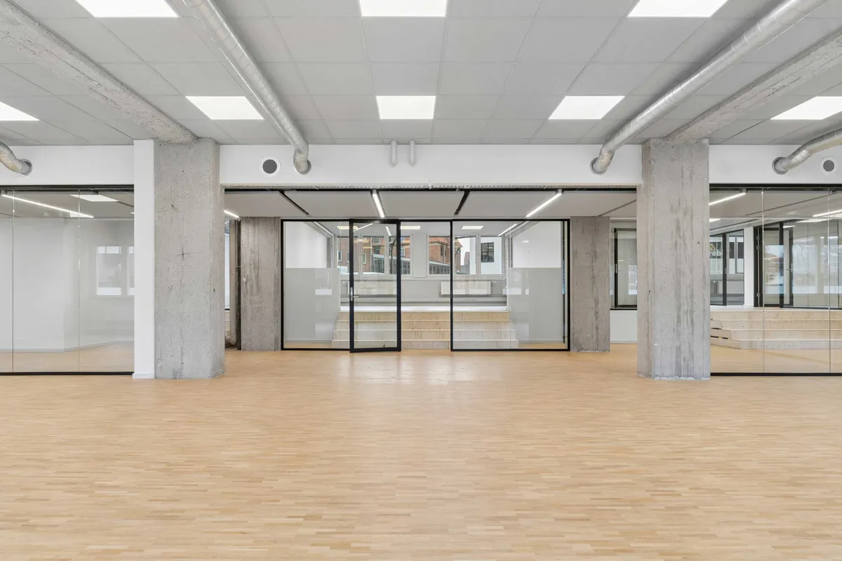 Et moderne kontor med store vinduer og lyse lofter samt trægulv. Større glasdøre med mørkere rammer og hvidt interiør.