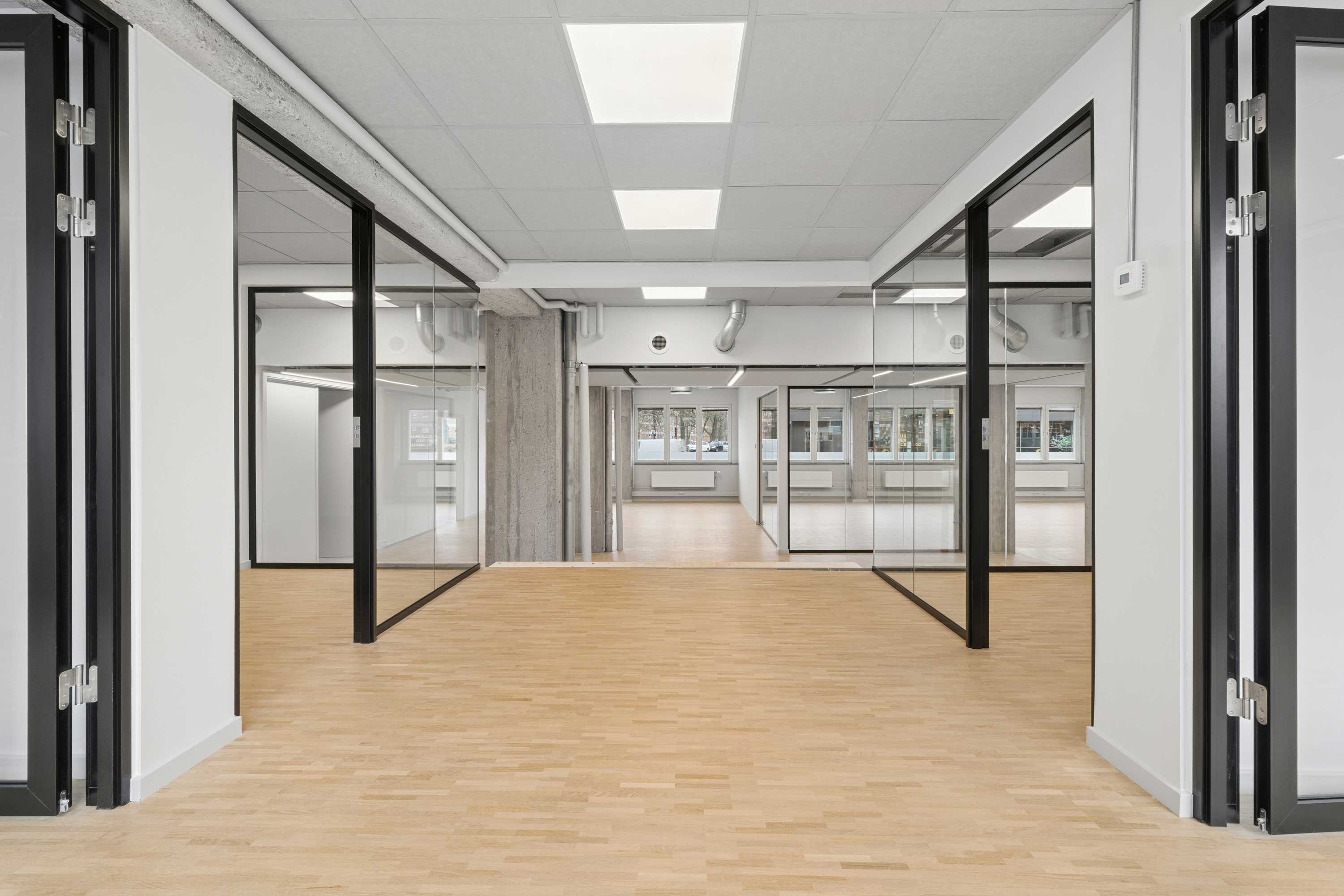 Stort, lyst kontor med glasvægge og hårdt trægulv. Lyse loftslamper og lyse, store vinduer. Sorte døre og gulve med hvide vægge.