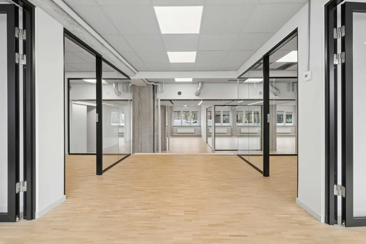 Stort, lyst kontor med glasvægge og hårdt trægulv. Lyse loftslamper og lyse, store vinduer. Sorte døre og gulve med hvide vægge.