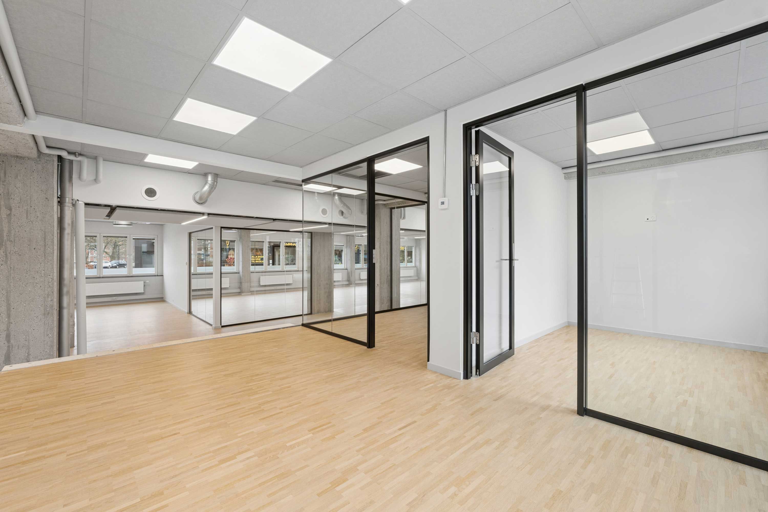 En tom kontorrum med hvidt loft og vægge samt glasdøre og -vinduer. Gulvet er lavet af træ og der er flere vinduer med udsigt til gadebelysningen