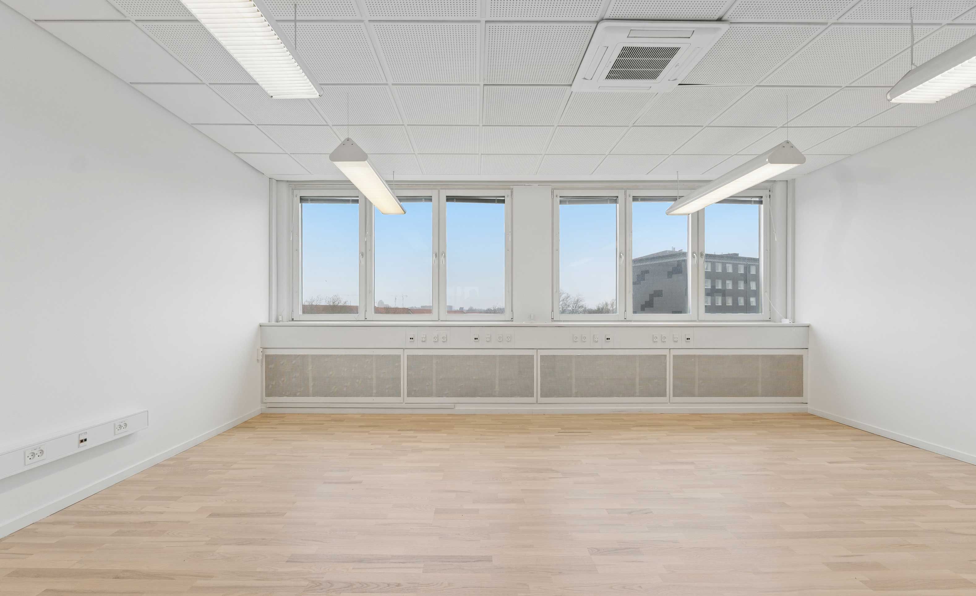 Tomt kontor med hvide vægge og loft, lysekroner og vinduer, lysegult trægulv og gule og hvide stikkontakter samt rullegardiner