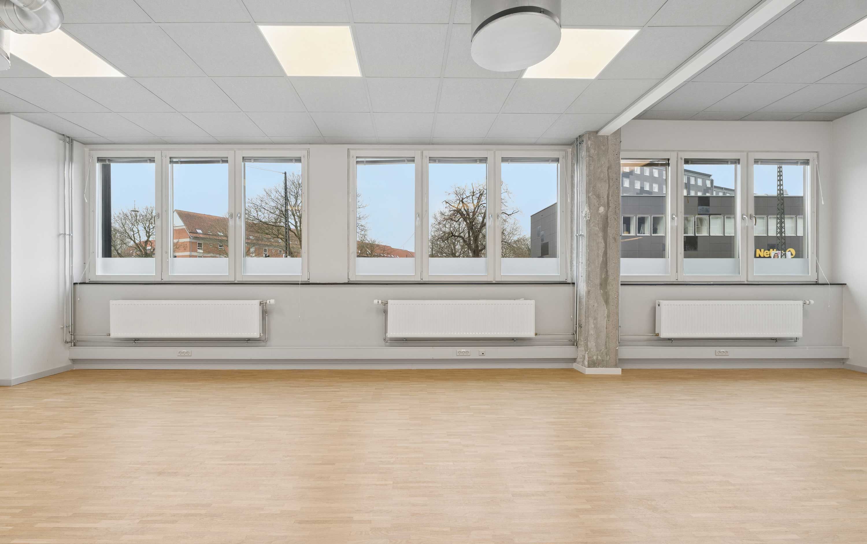 Kontor i Rønnegade med lyseruder og højt loft. Vinduerne vender mod skov og bygninger. Gulvet er lavet af træ, og der er radiatorer og lys i loftet.