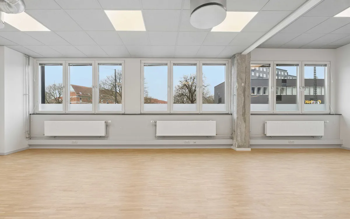Kontor i Rønnegade med lyseruder og højt loft. Vinduerne vender mod skov og bygninger. Gulvet er lavet af træ, og der er radiatorer og lys i loftet.