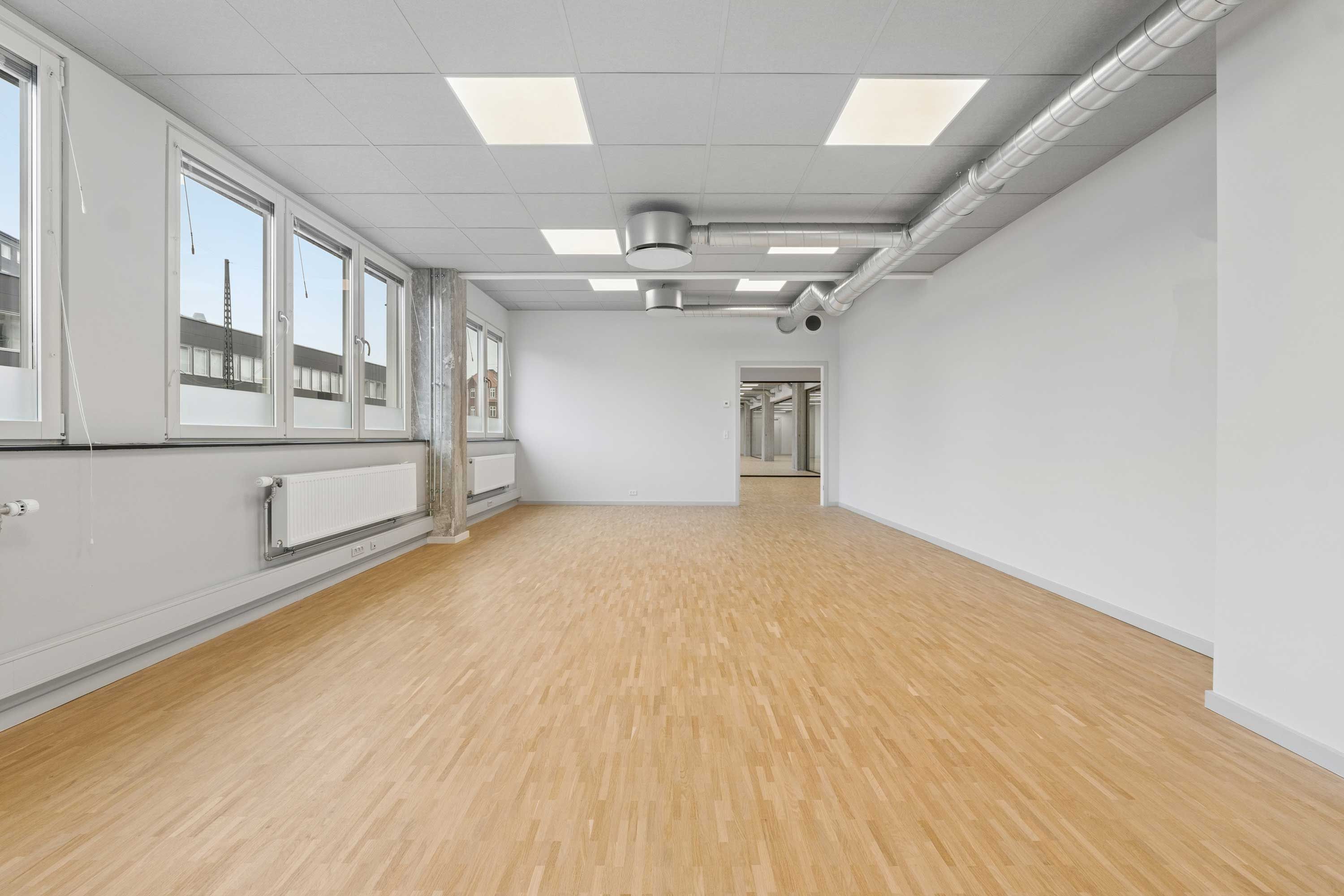 Bredt kontor med hårdt trægulv og store vinduer. Hvidt loft med lysekroner og ventiler. Væggene er hvide og renset. Der er radiatorer og stikkontakter.