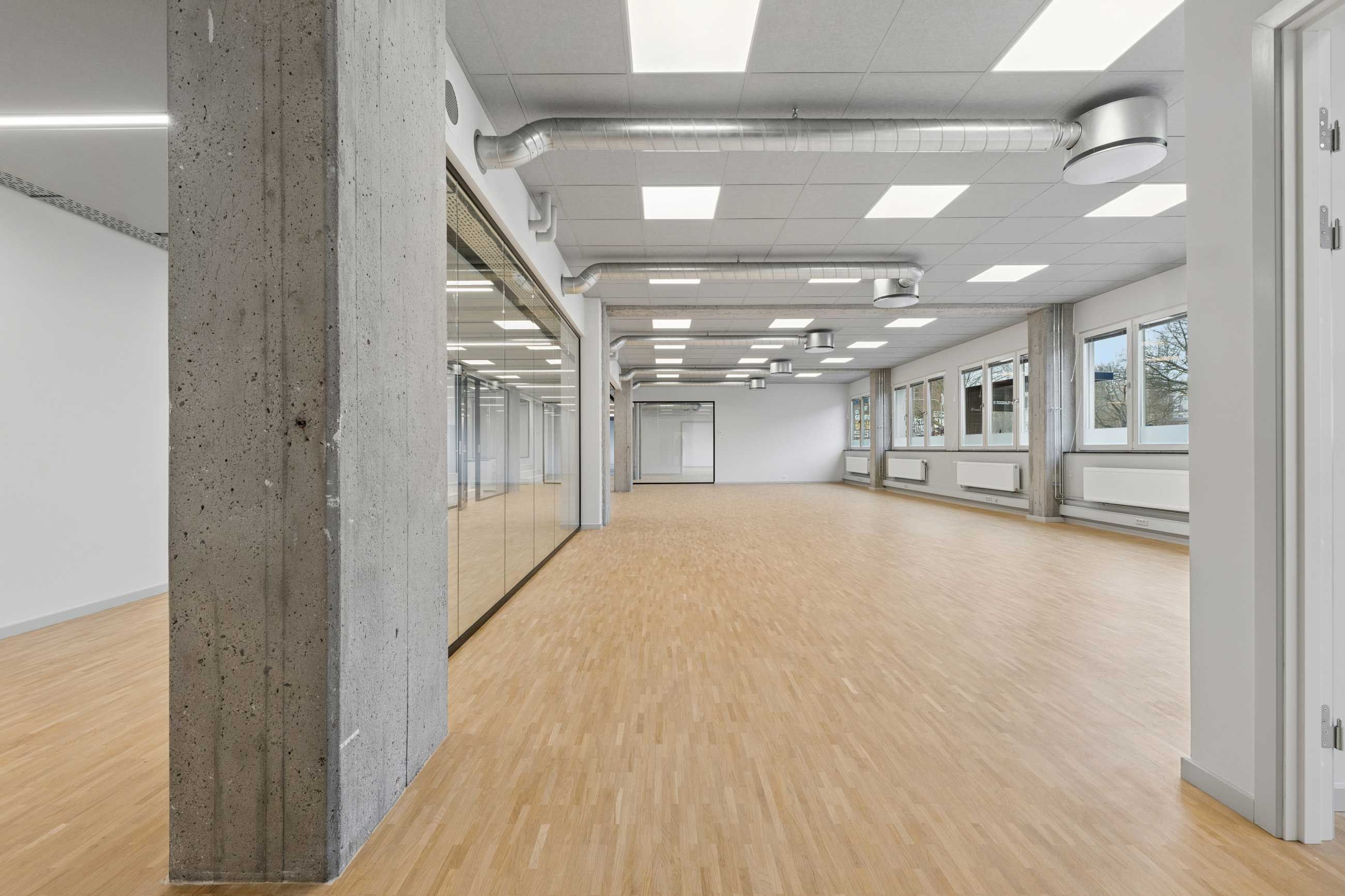 Et sammenhængende kontor med lyse loftslampe, glasvæg, hvid væg og massiv trægulv. Et stort betonbælte står mod væggen. Rummet er ubeboet.