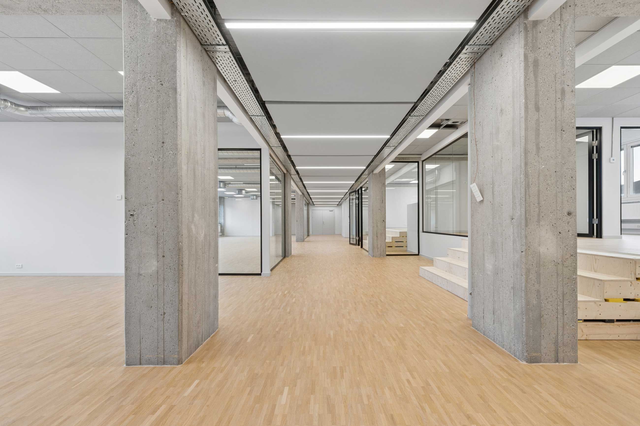 Kort hal med lyse trægulve og høje konkrete søjler og loftspaneler. Vægge og gulve er hvide, og vinduer er i glas. Der er stige og trapper langs væggen