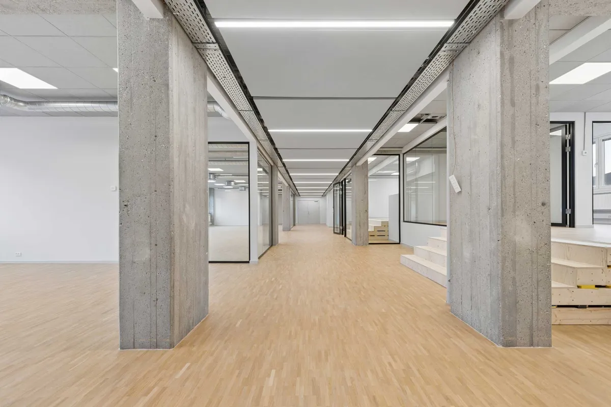 Kort hal med lyse trægulve og høje konkrete søjler og loftspaneler. Vægge og gulve er hvide, og vinduer er i glas. Der er stige og trapper langs væggen