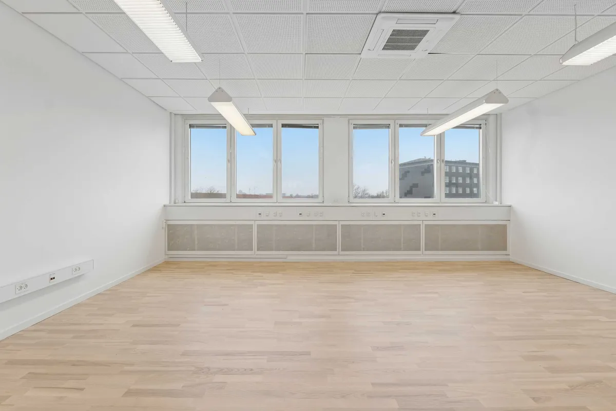 Lyst, moderne kontor med hvidt loft og vinduer, tæt på metro. Parketgulv, hvide vægge og flere stikkontakter. I baggrunden er der bygninger og et lyst himmelbånd.