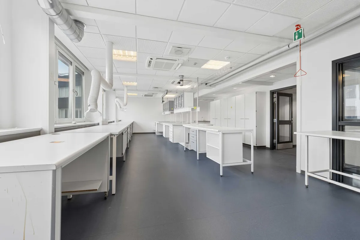 Modernt laboratorium med lyse vægge og højloft. Værelset er udstyret med hvide skriveborde og kabinetter. Luftkanaler og ventilation i loftet. Døre og vinduer er i hvidt og sort.