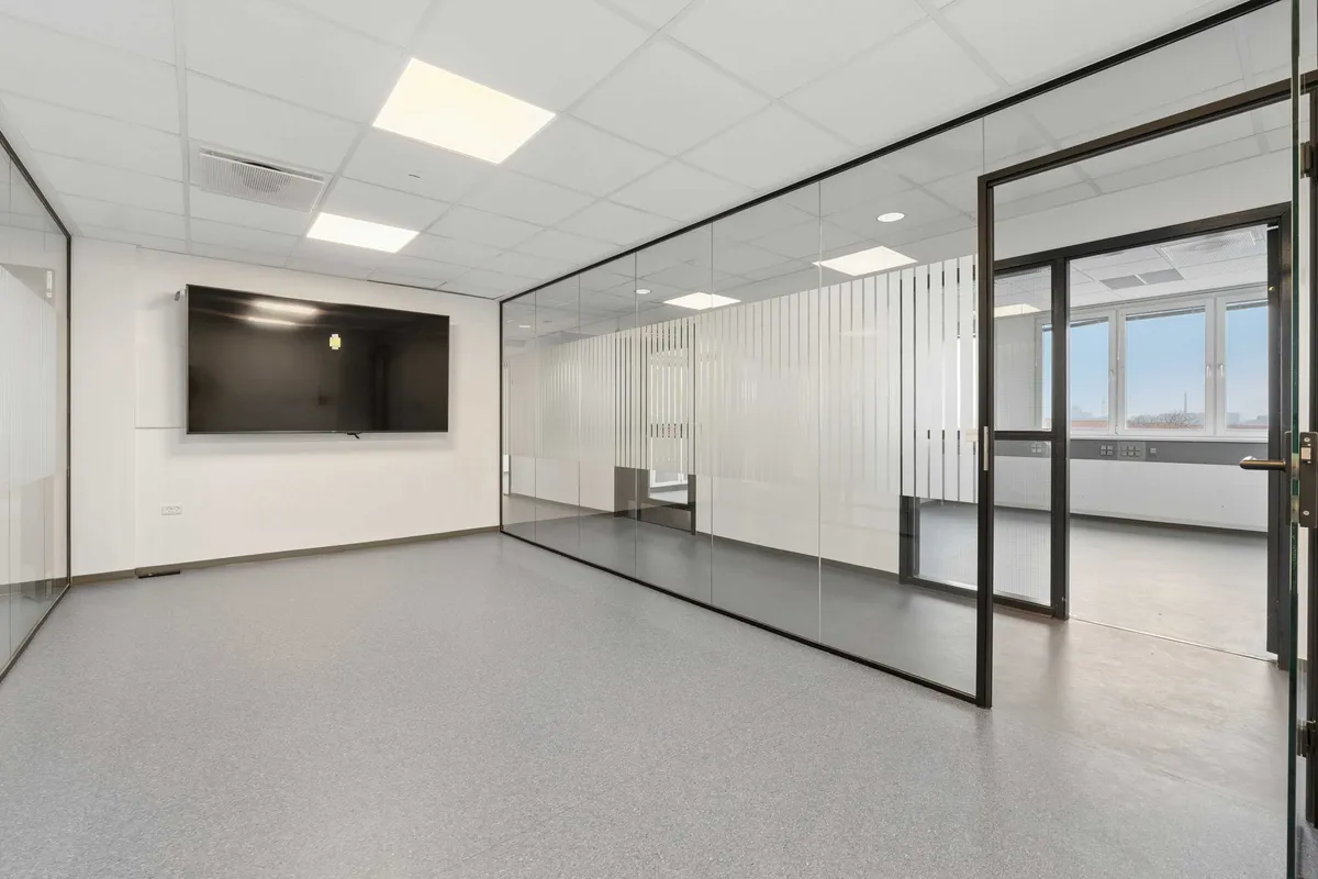 Modernt kontor med højt loft, glasvægge og lysegulve, hvor en stor tv-skærm er monteret på væggen. Glassdøre og vinduer giver masser af naturligt lys