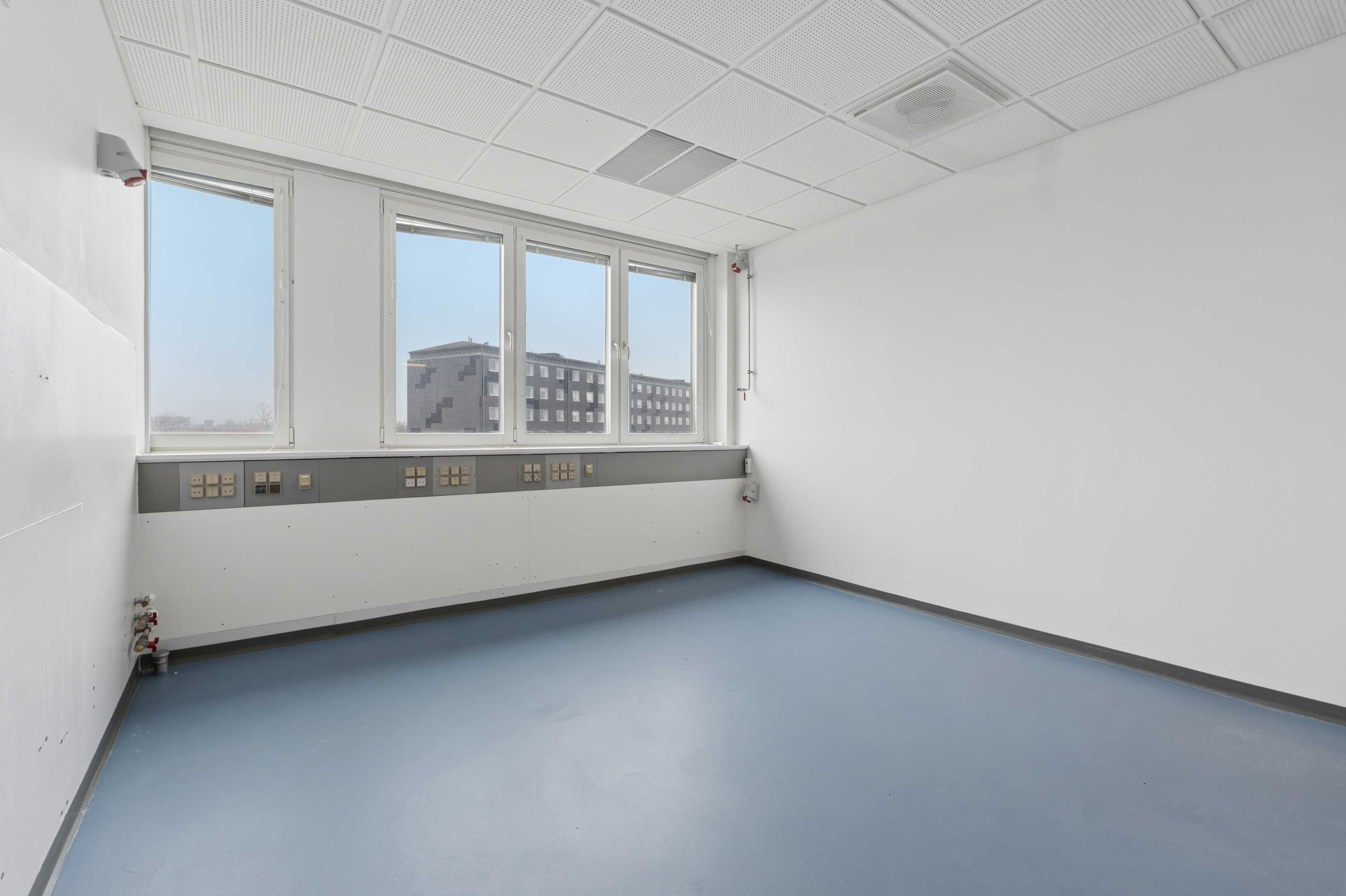 Moderne laboratorie med hvide vægge, blå gulv og tre store vinduer med udsigt til bygning. Gulvet er renset og der er stikkontakter og et ventilationssystem på væggen.
