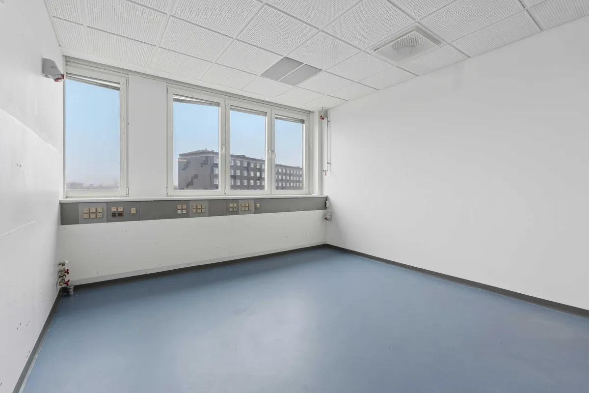 Moderne laboratorie med hvide vægge, blå gulv og tre store vinduer med udsigt til bygning. Gulvet er renset og der er stikkontakter og et ventilationssystem på væggen.