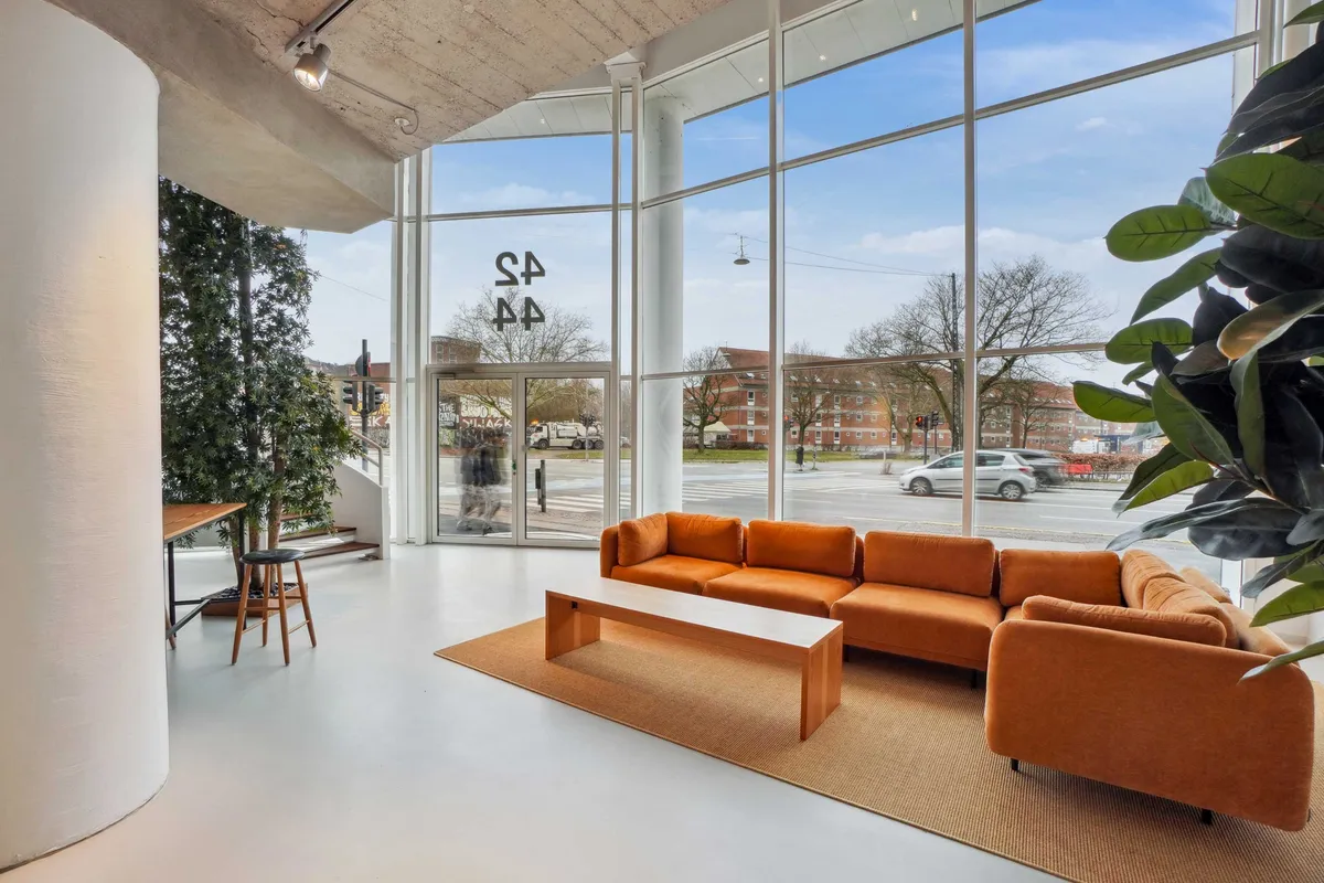 Lyst og moderne kontorrum med orange sofa, hvidt loft, lyse gulve og stort glasvindue med udsigt til byen, skriver "S44" på vinduet og lysekroner