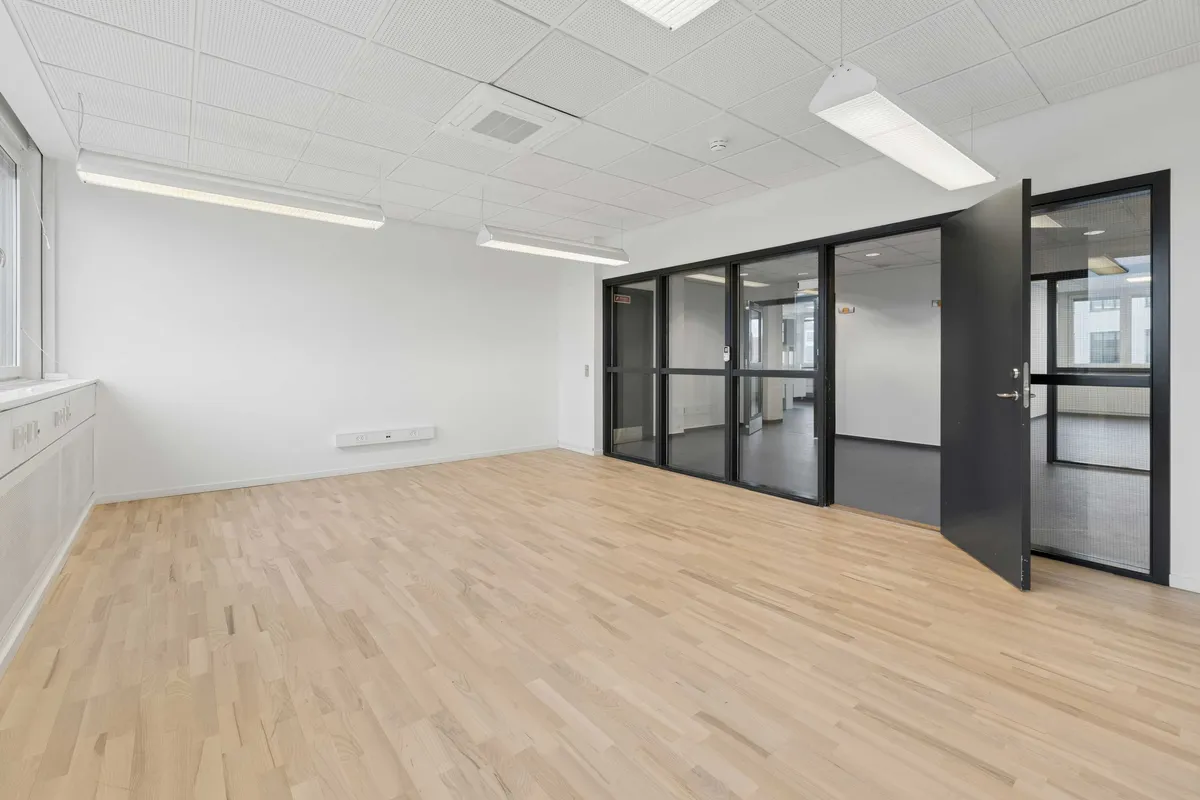 Moderne kontor med højloft og lyst indre. Værelse med højt gulv, glasdøre og hvide vægge. En dør er åben, og der er lysekroner og luftkanaler i loftet.