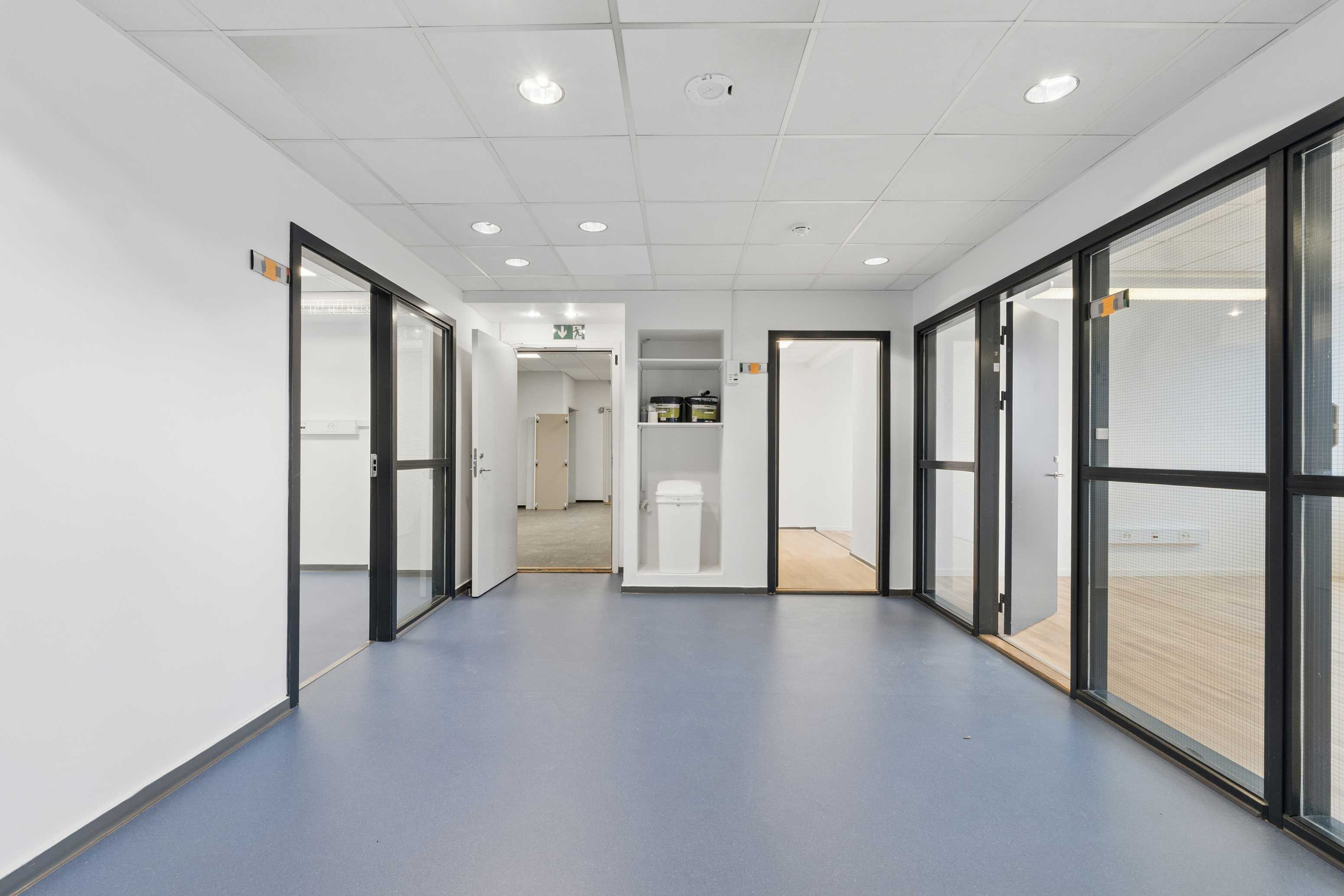 Lyst og moderne kontor med åbne døre, glasruder og lyse loftslampe. Gulvbelægning og lyse vægge. Et hvidt skab med tøjkurv i det ene værelse