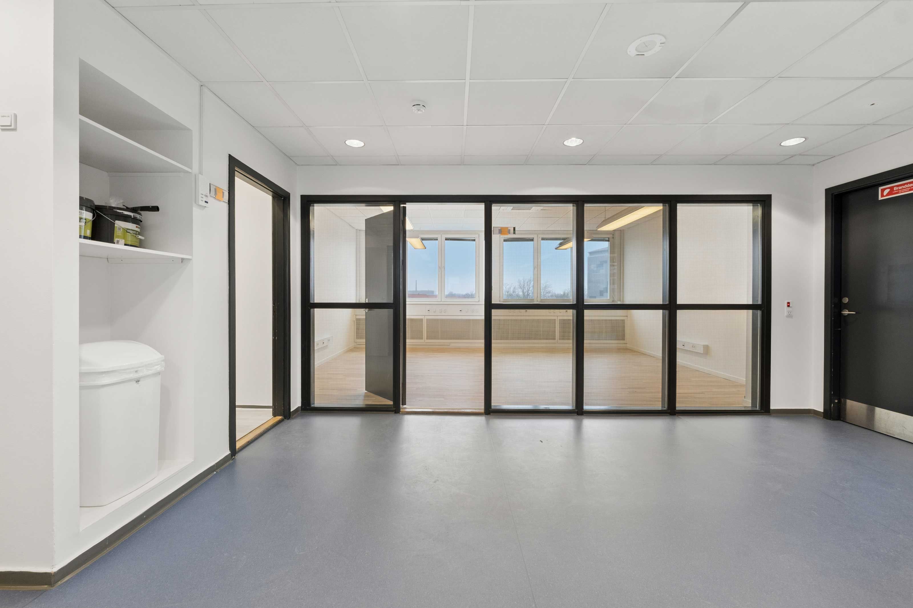 En stor og lys laboratorielokation med glasruder og indgangsdøre i lyse nuancer. Loftet er hvidt og renset. Væggene er hvide, og der er et skab med beholdere på.