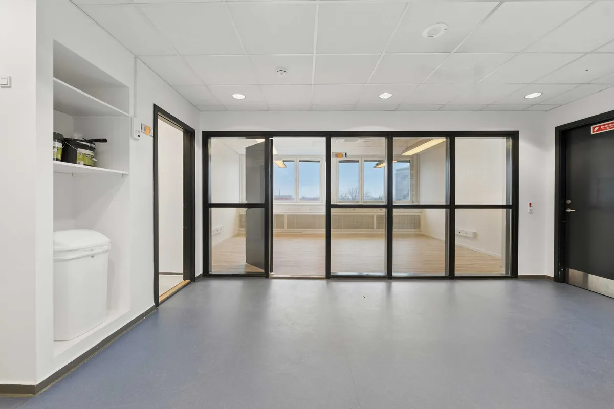 En stor og lys laboratorielokation med glasruder og indgangsdøre i lyse nuancer. Loftet er hvidt og renset. Væggene er hvide, og der er et skab med beholdere på.