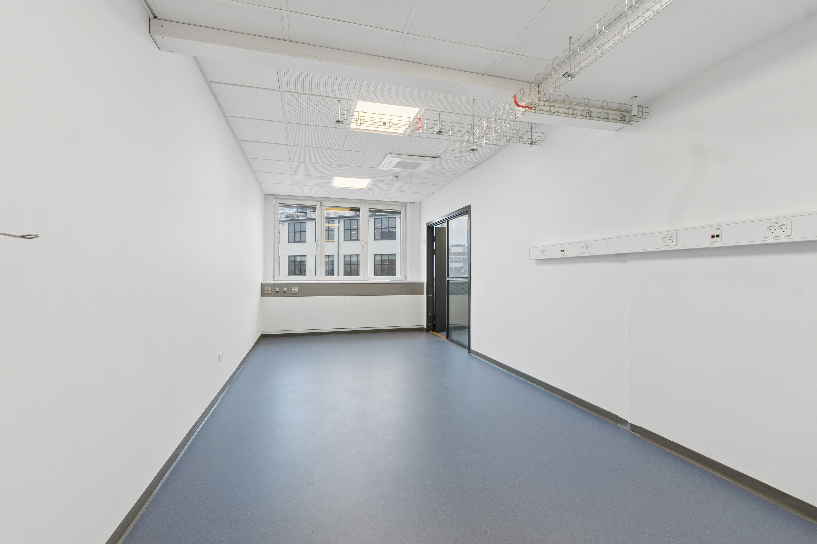 Lyst laboratorium med hvidt loft og gulv samt to døre i hvide vægge. En række elstik og udstik er monteret på væggen. Et vindue vender ud mod en bygning med mange vinduer