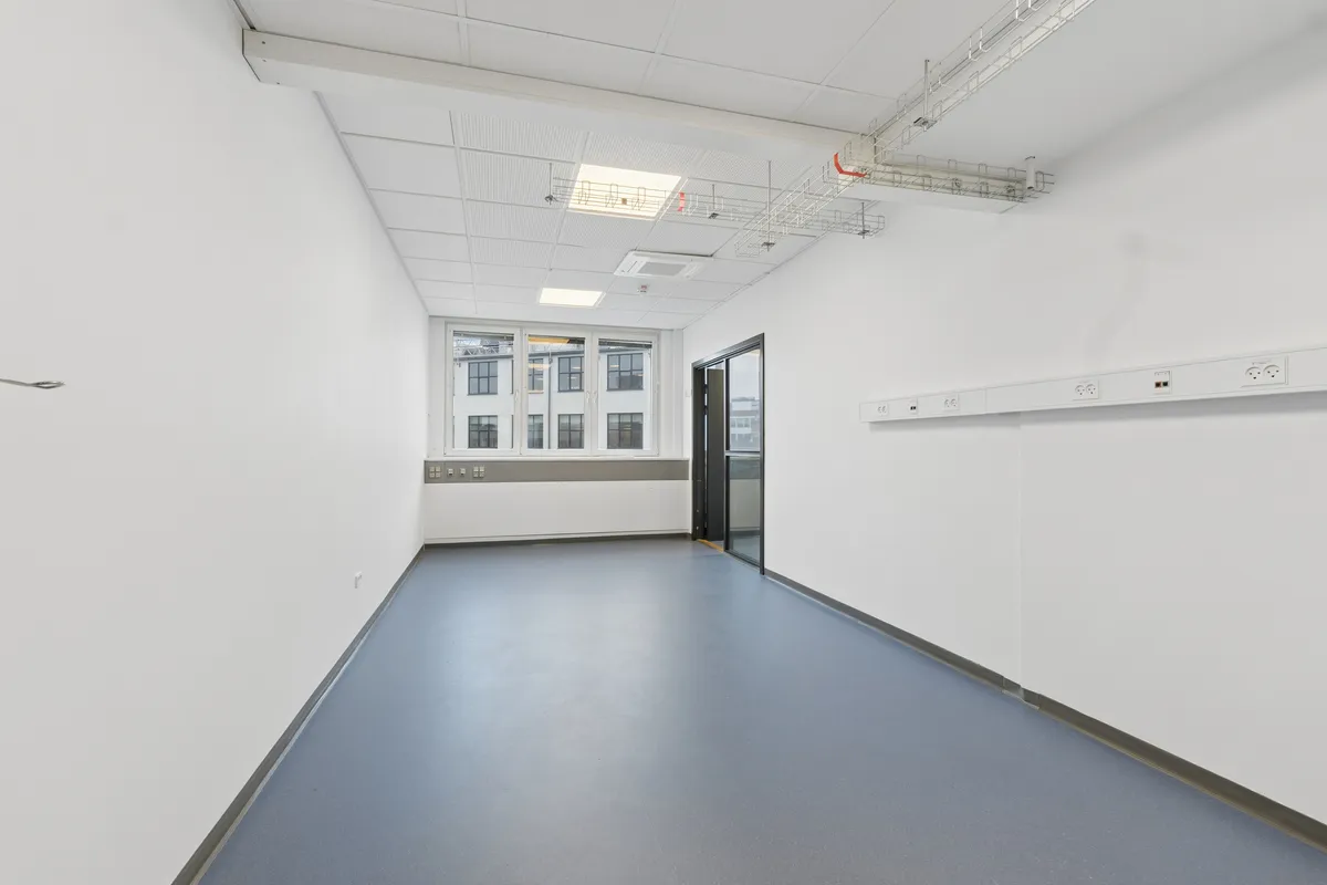 Lyst laboratorium med hvidt loft og gulv samt to døre i hvide vægge. En række elstik og udstik er monteret på væggen. Et vindue vender ud mod en bygning med mange vinduer