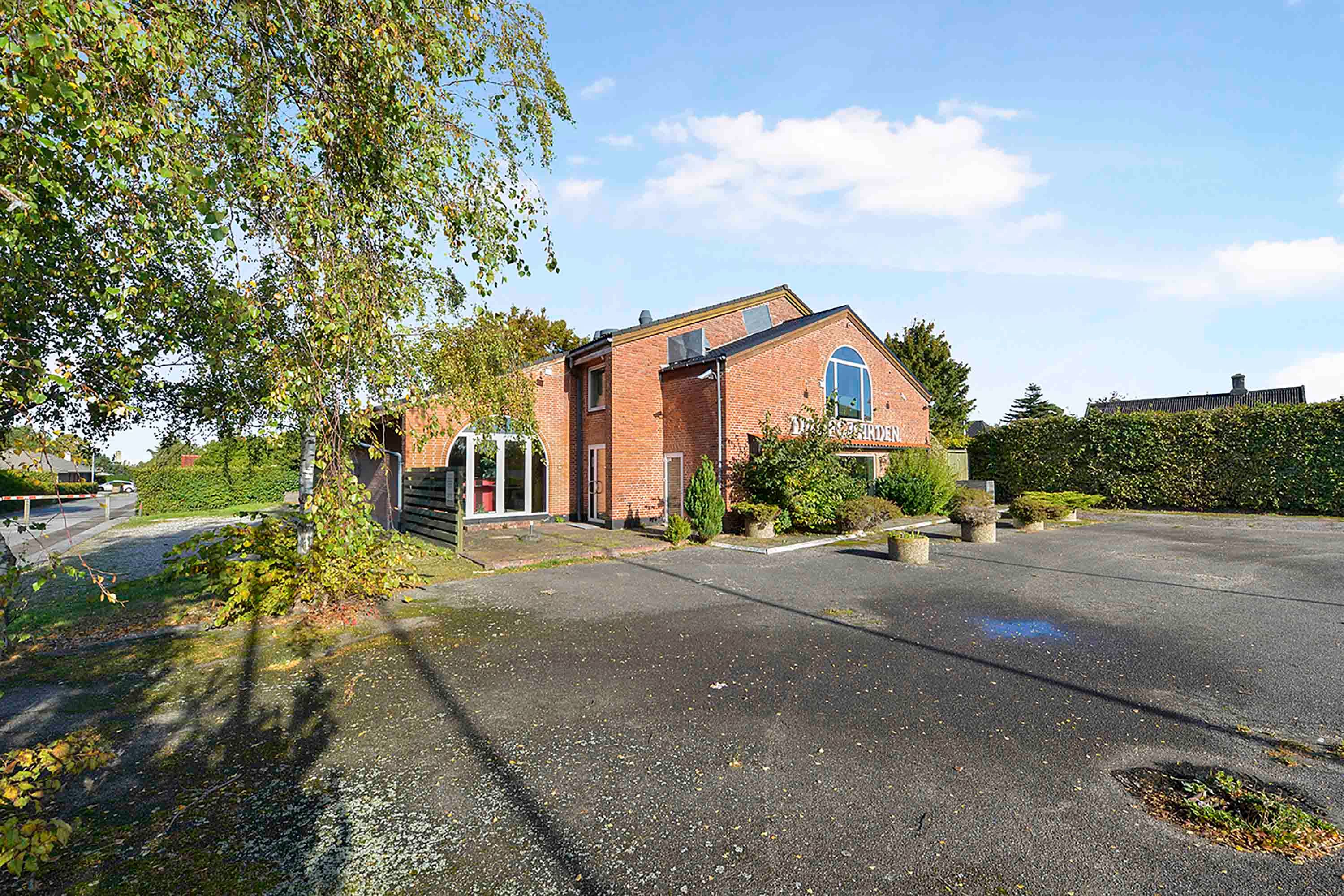 Restaurant i Greve med terrasse og parkeringsplads med buske og træer omkring. Et rødmurhus med glasruder og solcellepaneler på taget