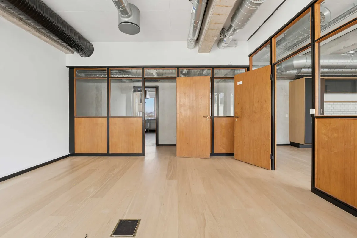 En stor åben stue med lyse gulve, to åbne døre og høje vinduer. En høj pipe løber langs loftet og gulvet er snydt med en grå stribe.