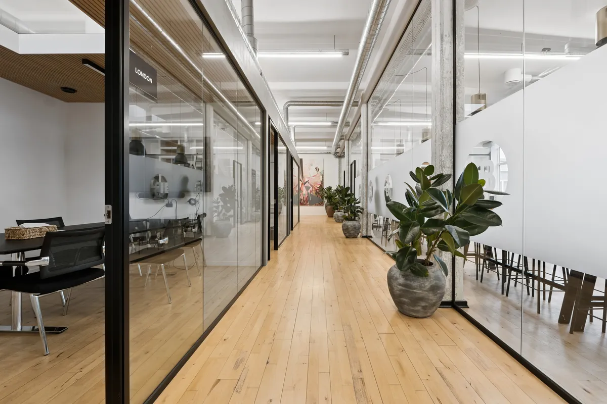 Glaspartier og møbler i en stor kontorhall med lyse trægulve og masser af grønne planter. Lamper hængende fra loftet og en rund spejl i væggen
