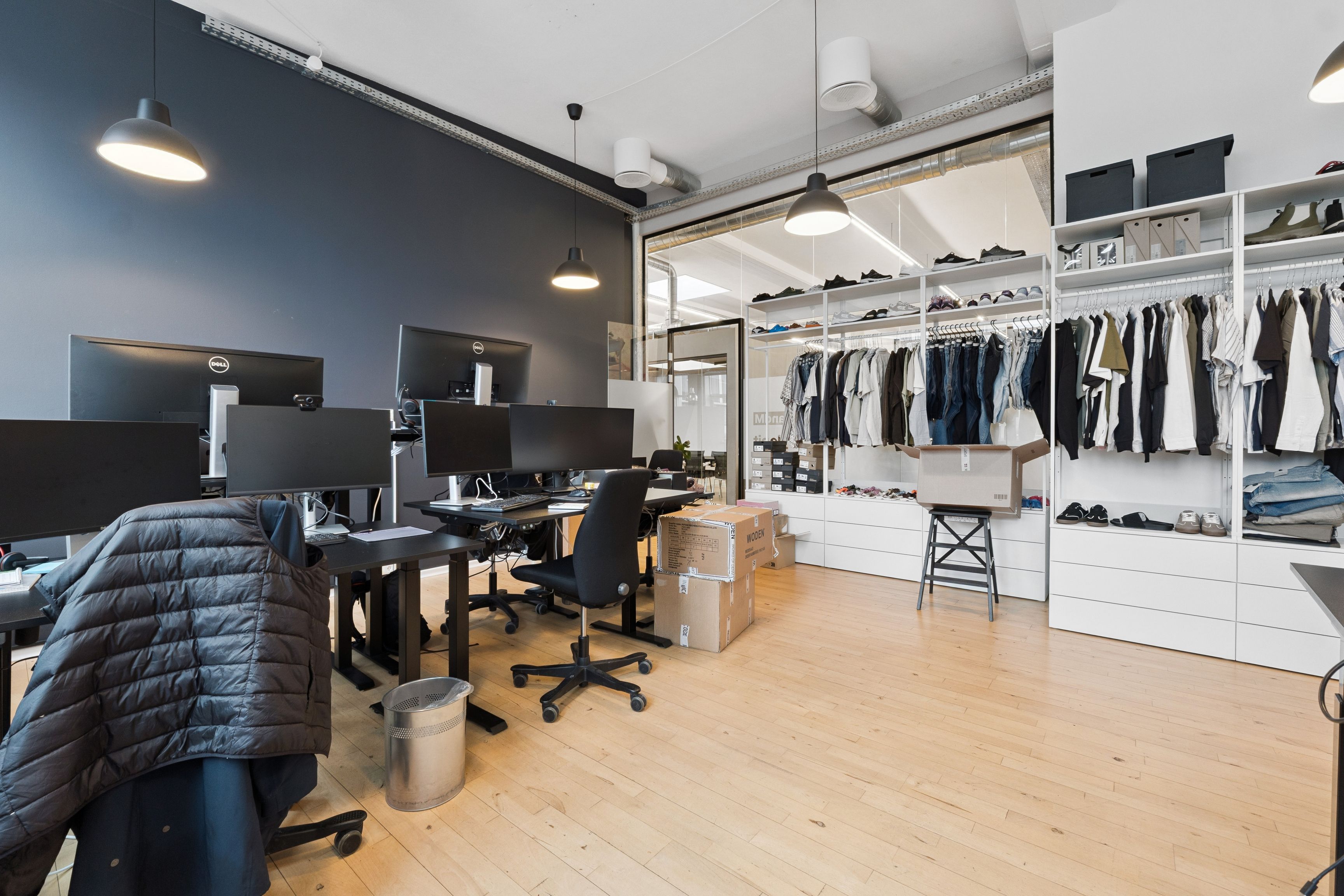 Kontorrum med skriveborde og skærme samt tøjskabe og kasser med tøj. Loftslampe, lyse og trægulv i rummet