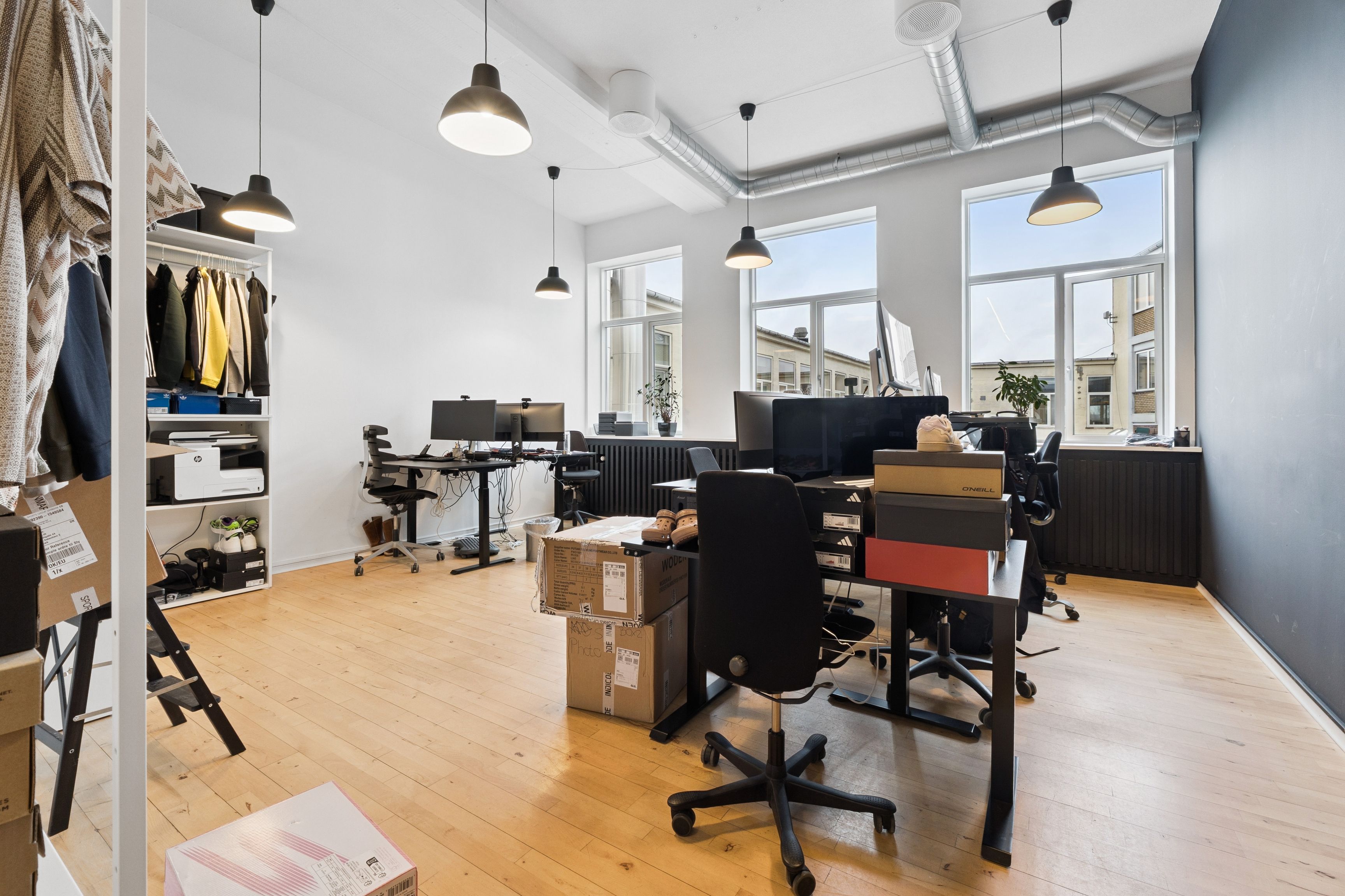 Kontor med moderne møbler og skriveborde, lyse loftslamper, vinduer og trægulv. Fladt loft og industrielle elementer som røre og plante.