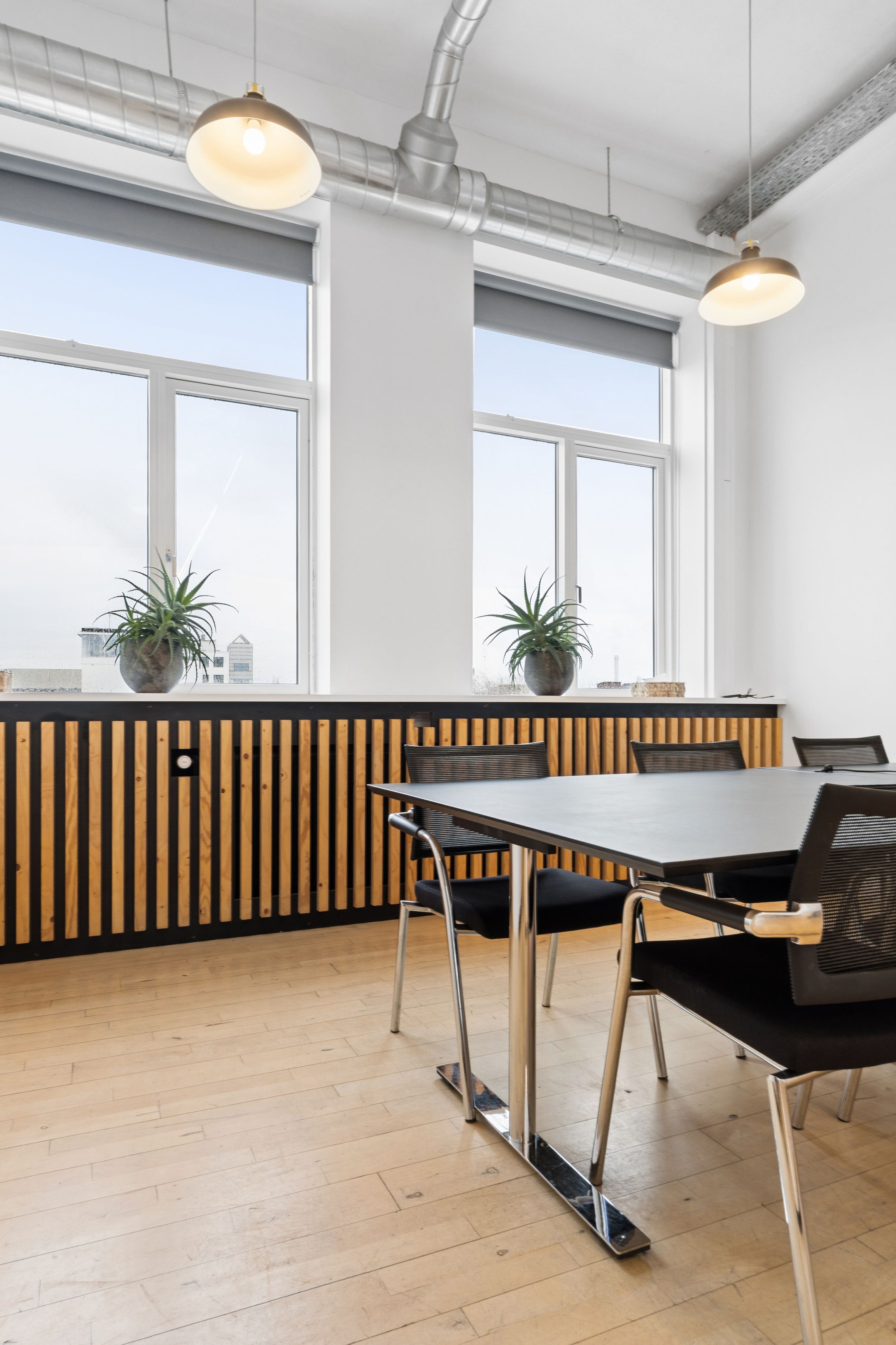 En tom, moderne konferencesal med store vinduer og lyse loftslamper. En stor, sort mødesalbord med fire moderne stole, træpaneler med plantepotter og luftkanaler i loftet