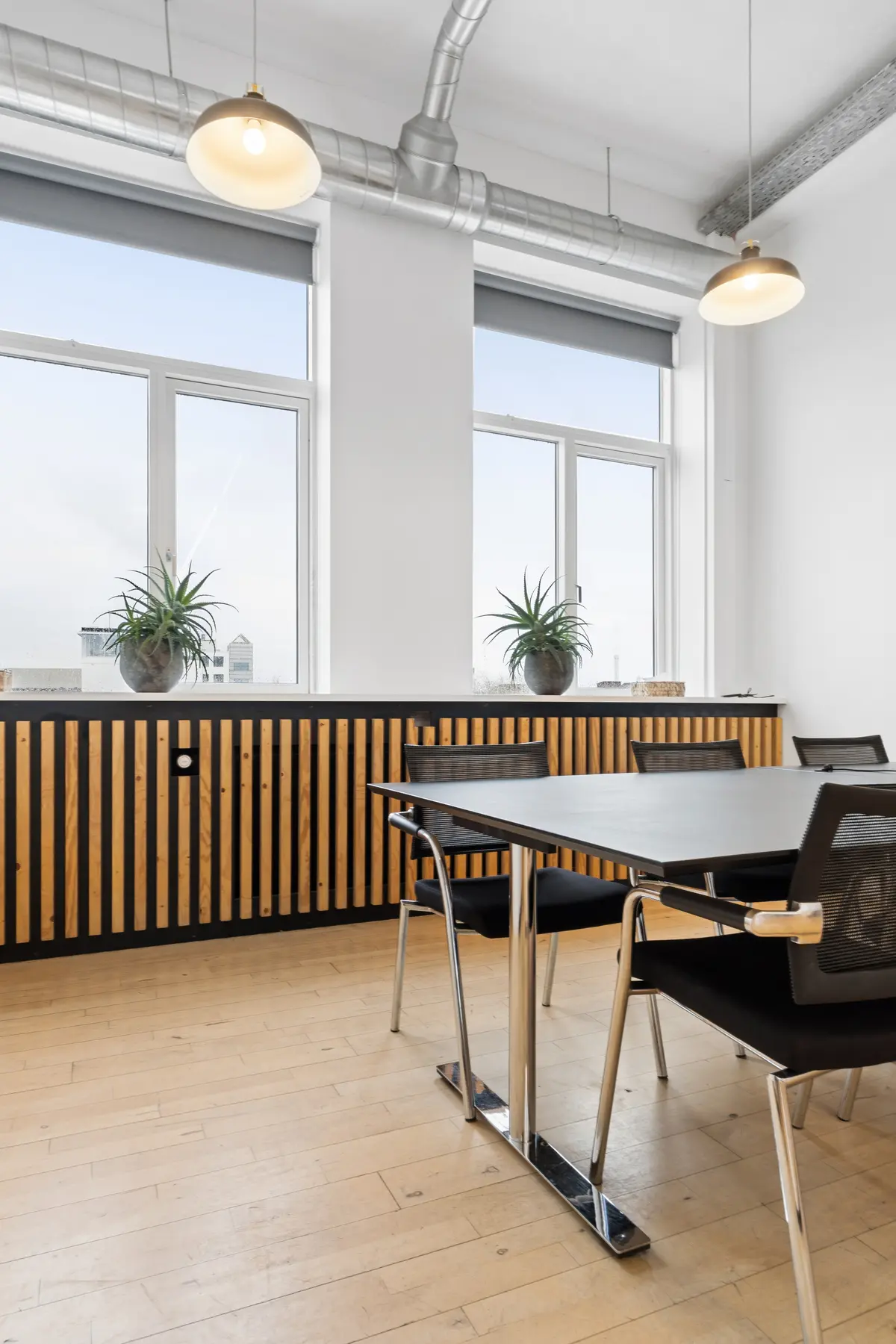 En tom, moderne konferencesal med store vinduer og lyse loftslamper. En stor, sort mødesalbord med fire moderne stole, træpaneler med plantepotter og luftkanaler i loftet