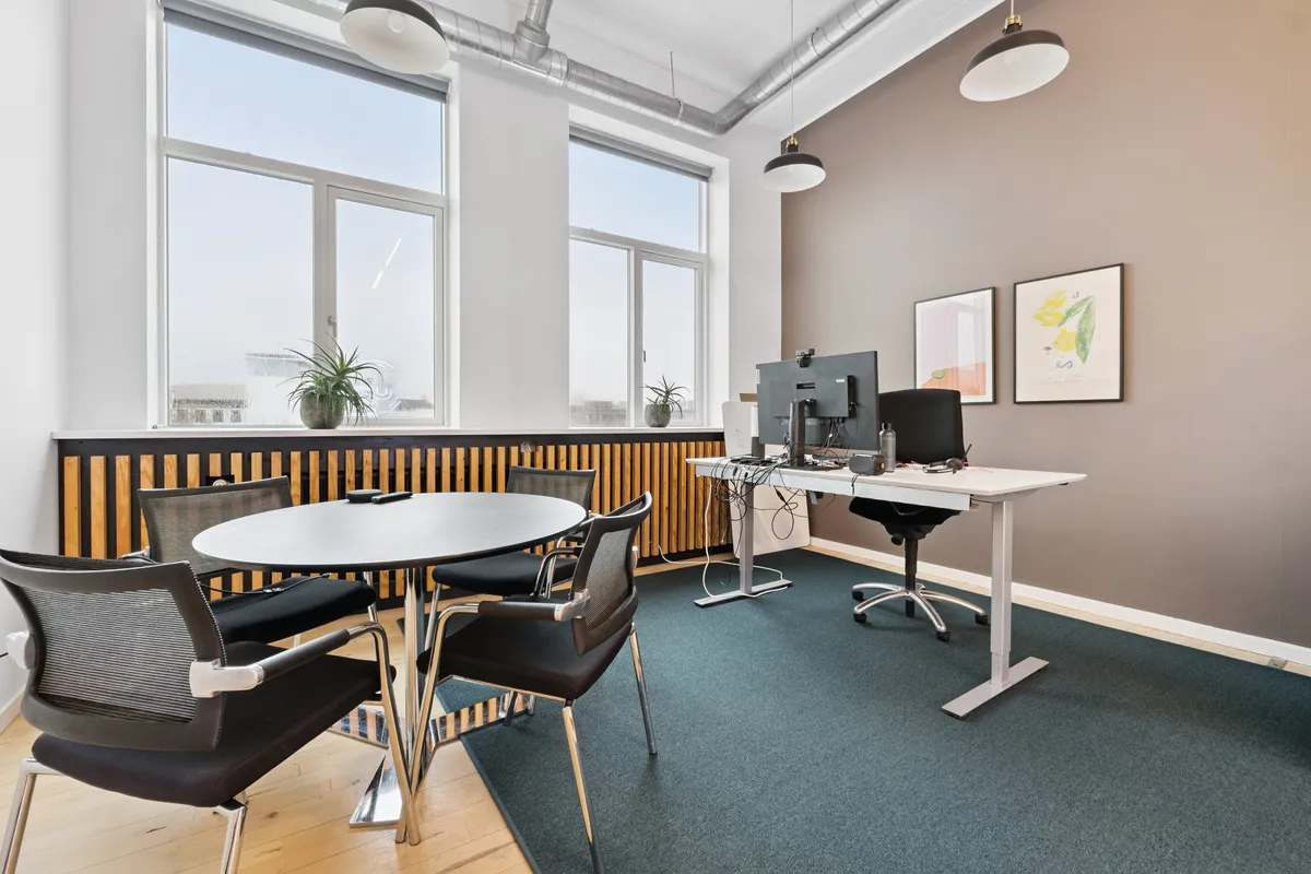 Kontor med lyse vinduer, lyse gulve, lyse loftslamper og lyse vægge. Kontorbord med to skærme og en stol. Bord med fire stole og en plante. To billeder hængende på væggen